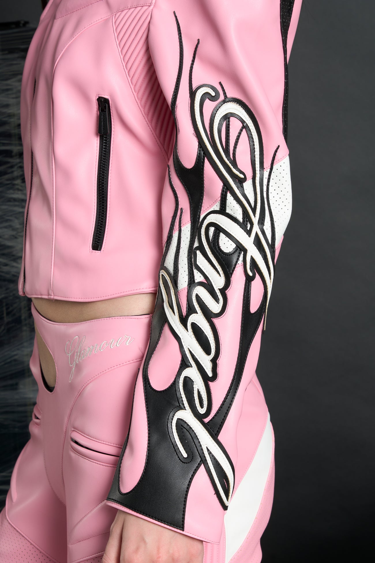 Model wears a pink motocross-style jacket made from vegan PU leather with flame topstitching, padded perforated paneling, topstiched sleeve slogan, ‘Angel’ embroidery on collar, and branded zippered front and pockets.