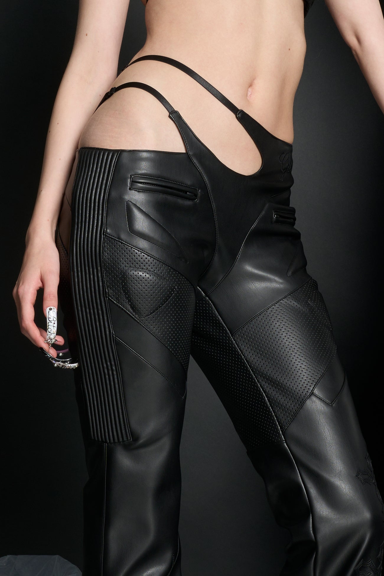 Model wears black vegan leather flare trousers with motocross paneling, padded details, flame and cross stitching, slogan embroidery, and an asymmetric waistband with adjustable elastic thong straps.