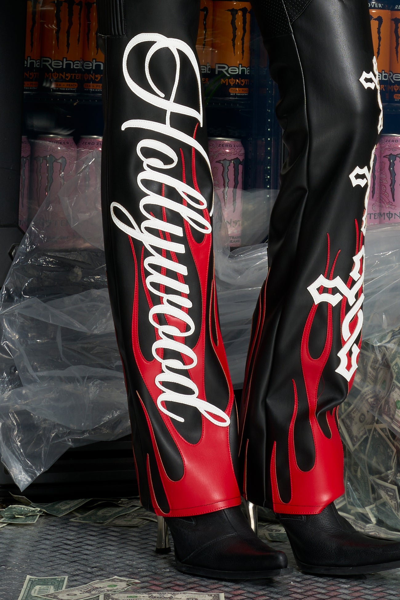 Model wears black/red vegan leather flare trousers with motocross paneling, padded details, flame and cross stitching, slogan embroidery, and an asymmetric waistband with adjustable elastic thong straps.
