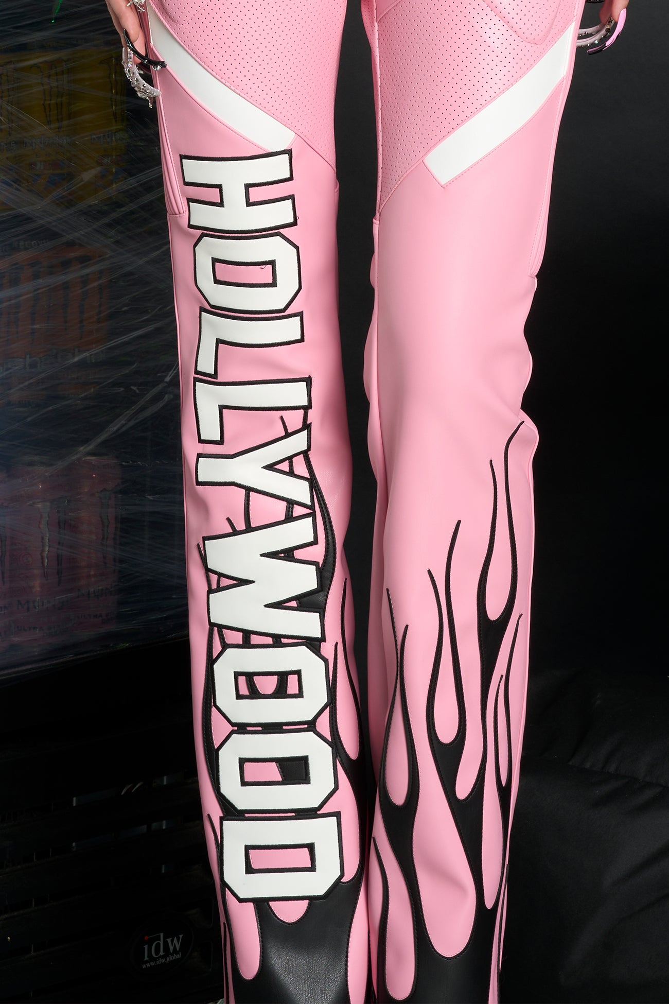 Model wears pink vegan leather flare trousers with motocross paneling, padded details, flame and cross stitching, slogan embroidery, and an asymmetric waistband with adjustable elastic thong straps.