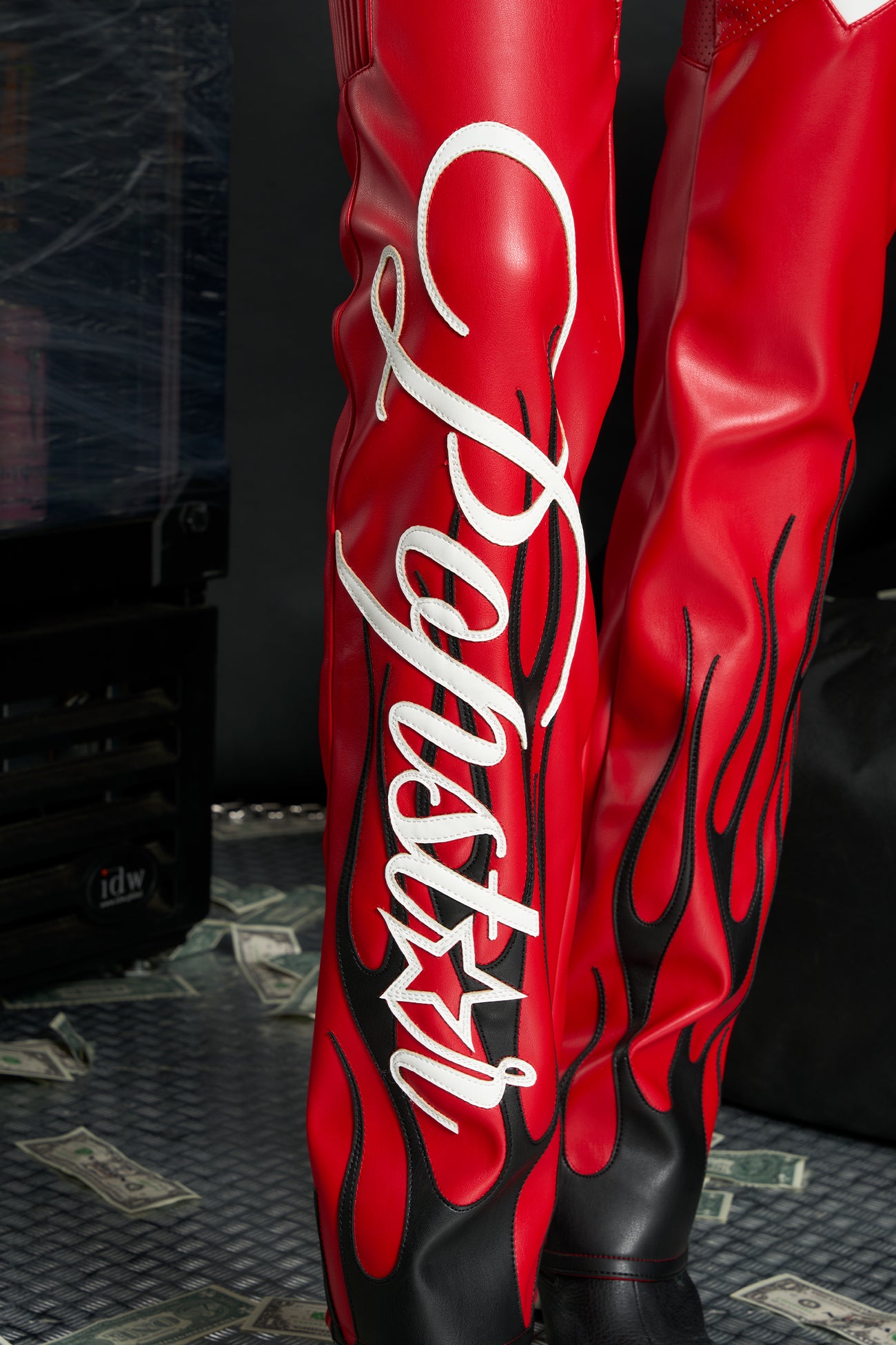 Model wears red vegan leather flare trousers with motocross paneling, padded details, flame and cross stitching, slogan embroidery, and an asymmetric waistband with adjustable elastic thong straps.