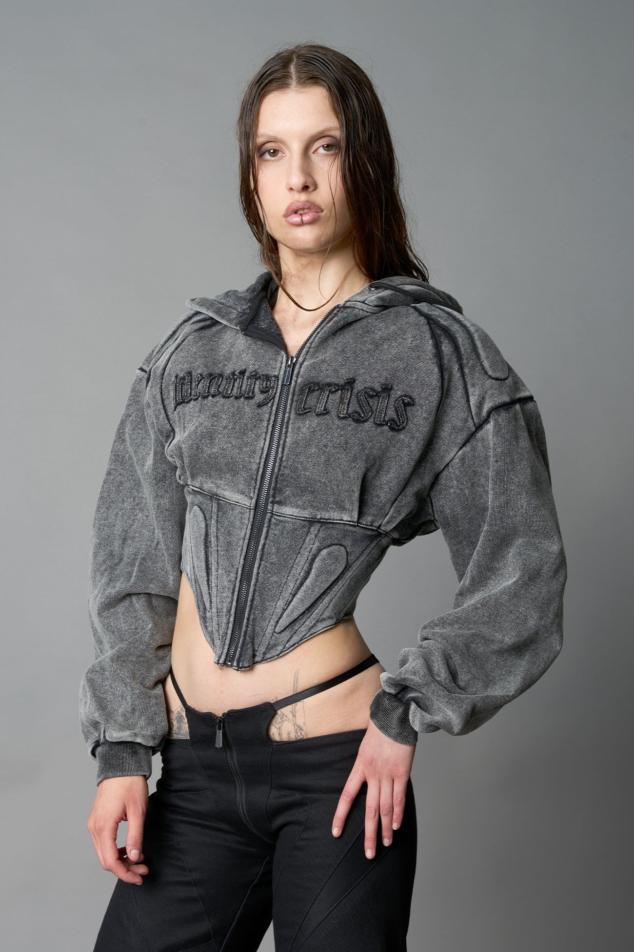 model in a washed-black cropped hoodie featuring structured corset-style paneling embroidered across the chest in tonal black