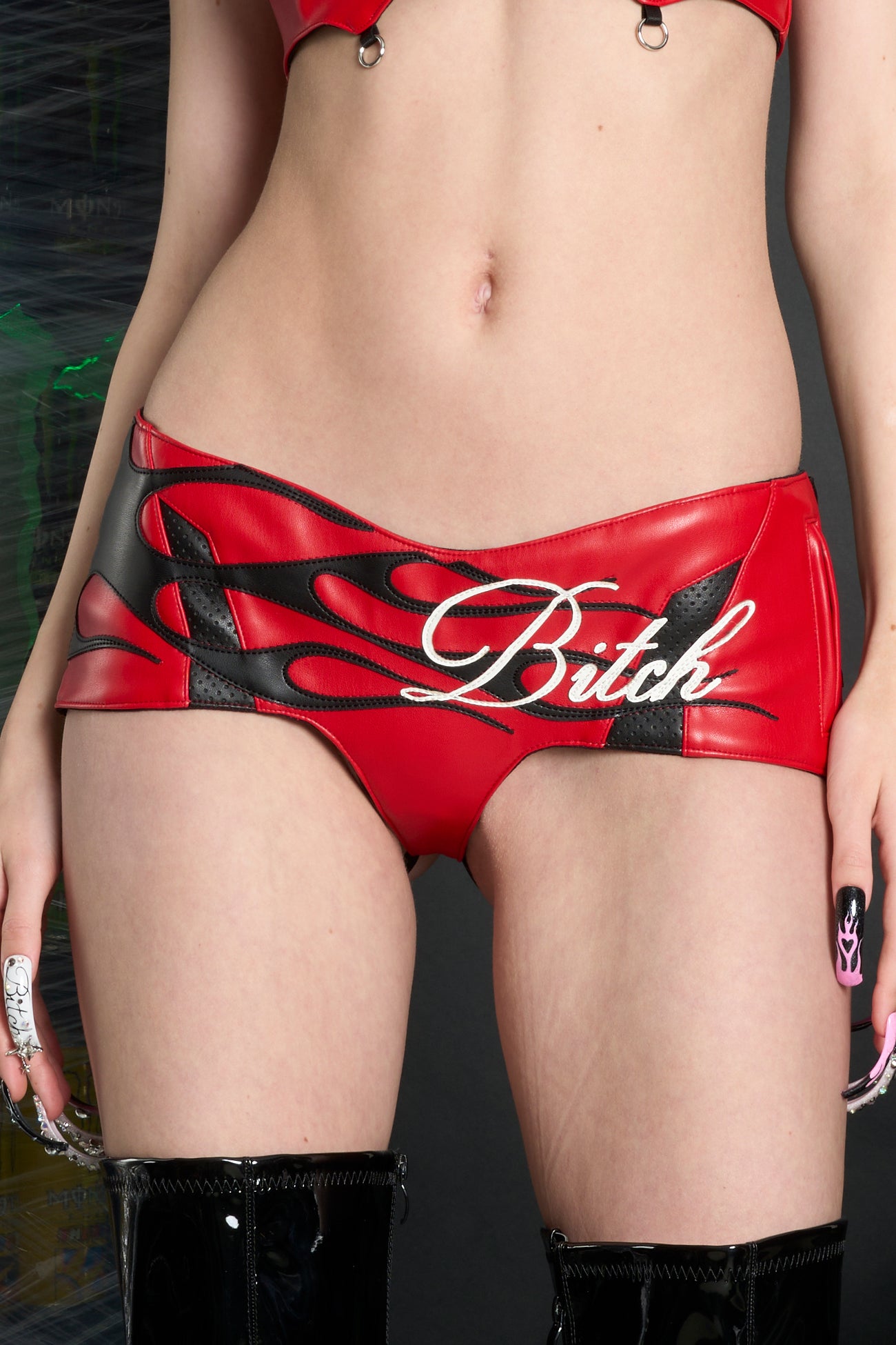 Model wears red low-rise vegan leather hot pants with adjustable elastic back straps. open back, flame topstitching, slogan topstitching