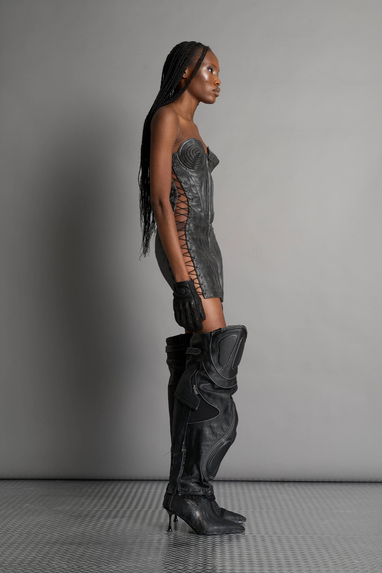 model wears the washed black vegan PU leather Namilia Moto Cone Dress, a strapless mini dress with cone cups, motocross panelling, branded zipper, side lacing, lycra back panel, and zippered breast pockets.