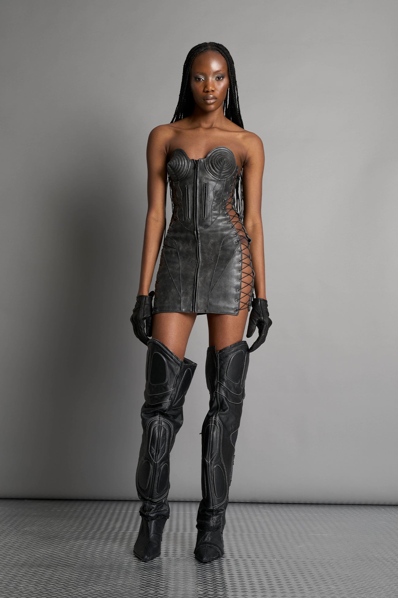 model wears the washed black vegan PU leather Namilia Moto Cone Dress, a strapless mini dress with cone cups, motocross panelling, branded zipper, side lacing, lycra back panel, and zippered breast pockets.