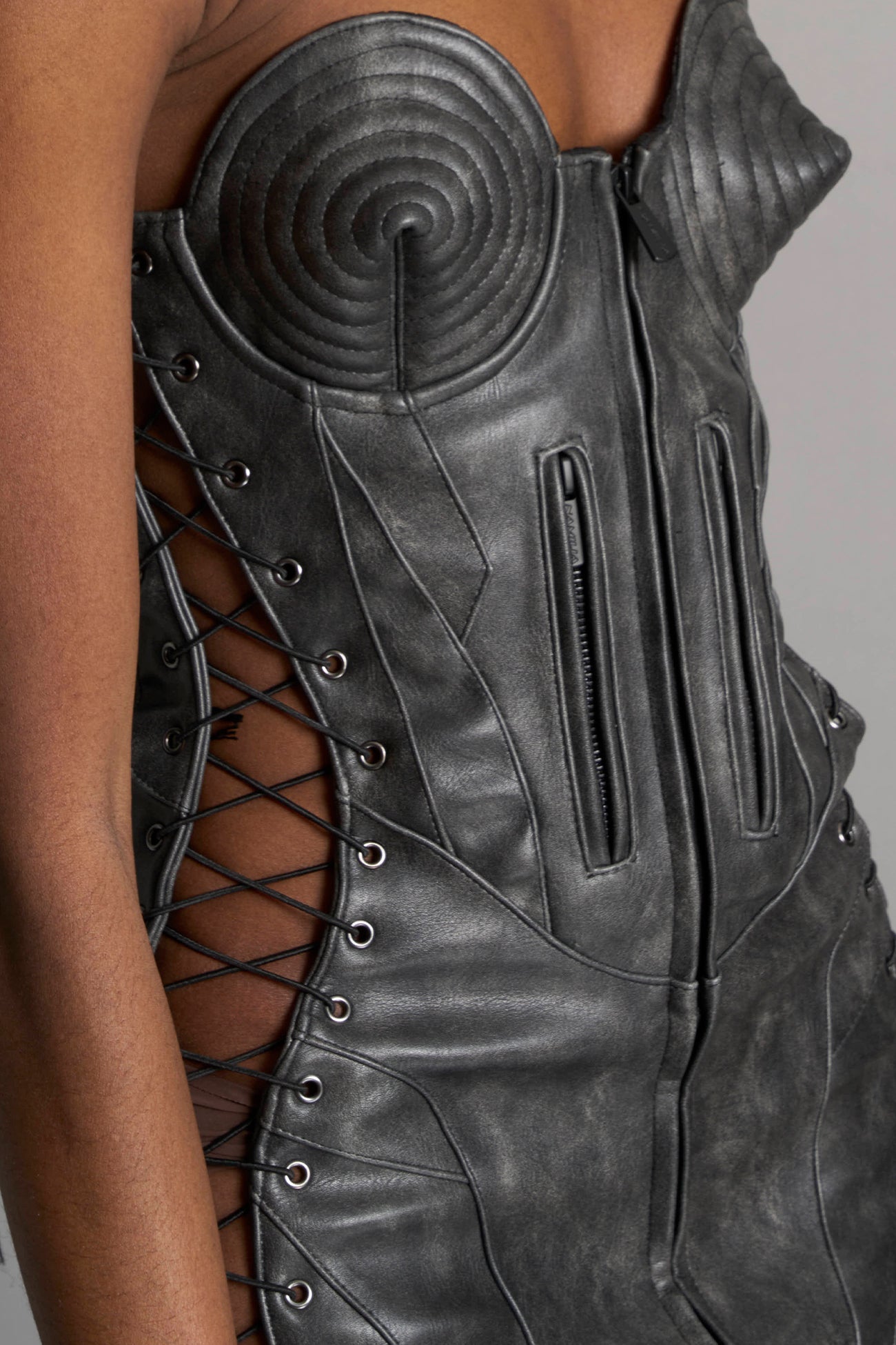 model wears the washed black vegan PU leather Namilia Moto Cone Dress, a strapless mini dress with cone cups, motocross panelling, branded zipper, side lacing, lycra back panel, and zippered breast pockets.