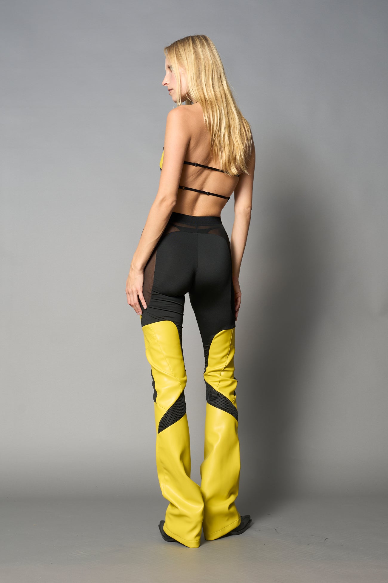 Model wearing high-waisted yellow vegan leather pants with mesh cutouts, moto-inspired paneling, and flared stacked legs – edgy streetwear trousers styled with chunky platform boots