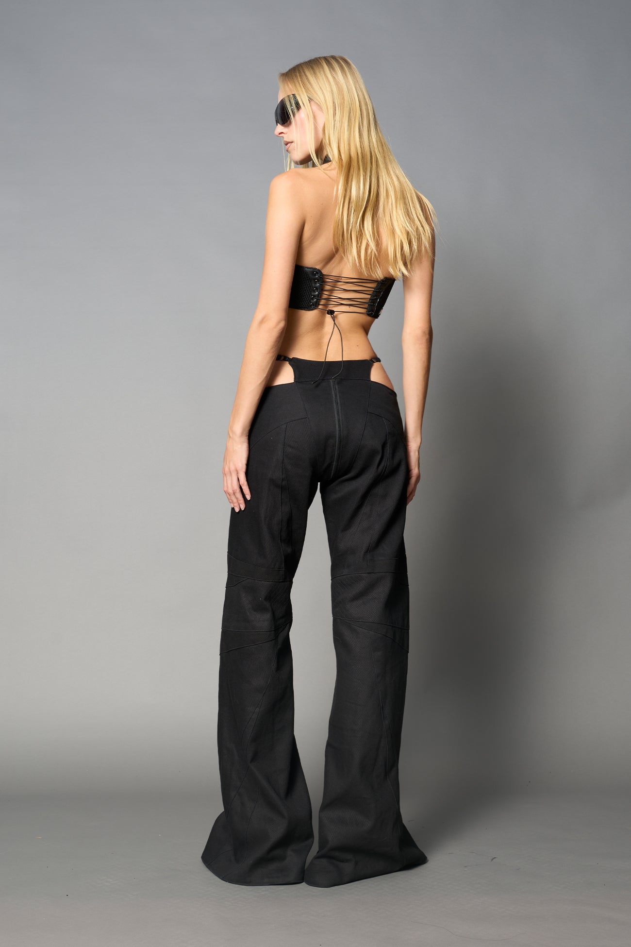 Low-rise flared pants in black cotton twill with bold side cut-outs and thong strap detailing. Featuring moto-style paneling, front zip closure, and a sculpted fit. A statement piece for rave, nightlife, and runway-inspired looks.