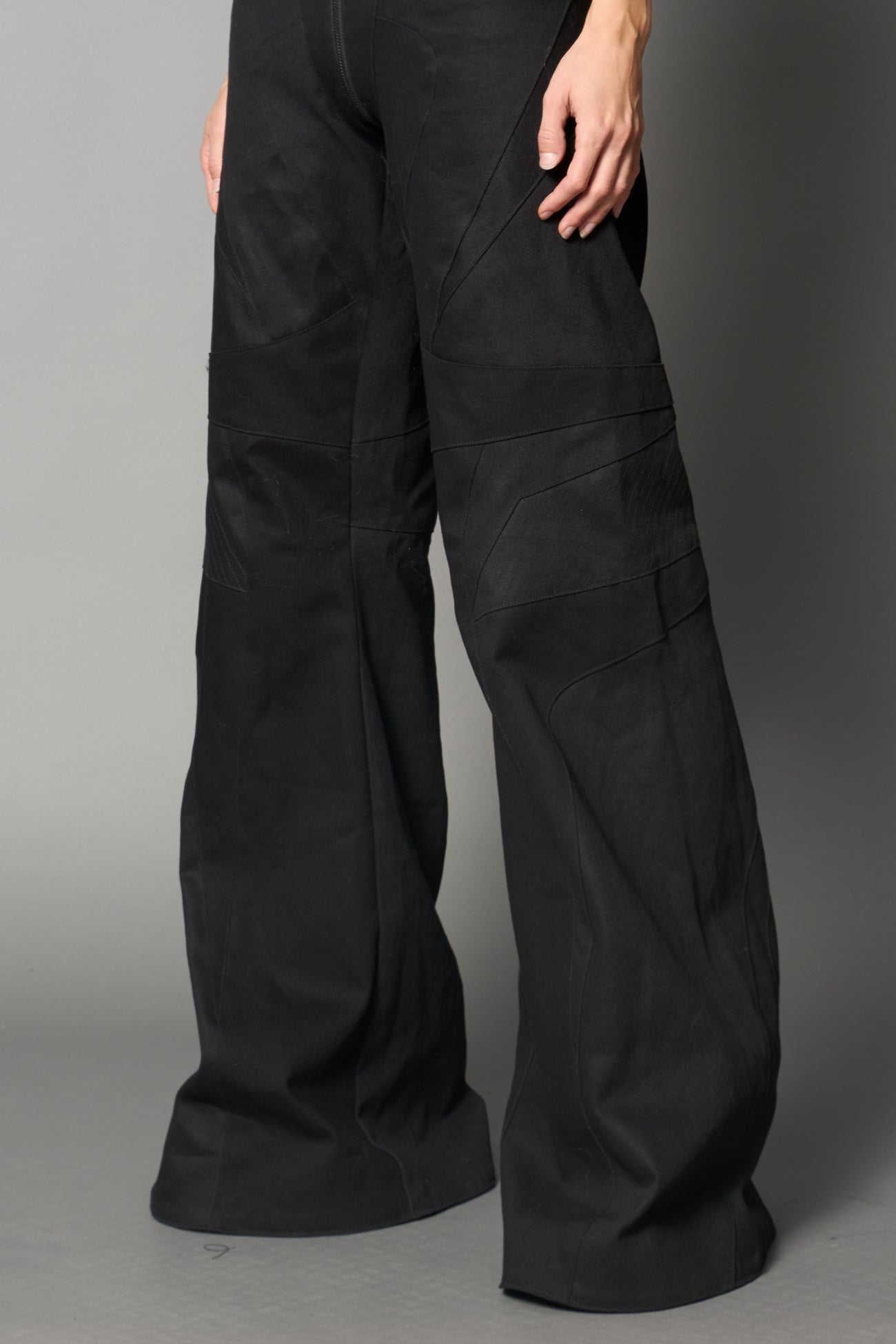 Low-rise flared pants in black cotton twill with bold side cut-outs and thong strap detailing. Featuring moto-style paneling, front zip closure, and a sculpted fit. A statement piece for rave, nightlife, and runway-inspired looks.