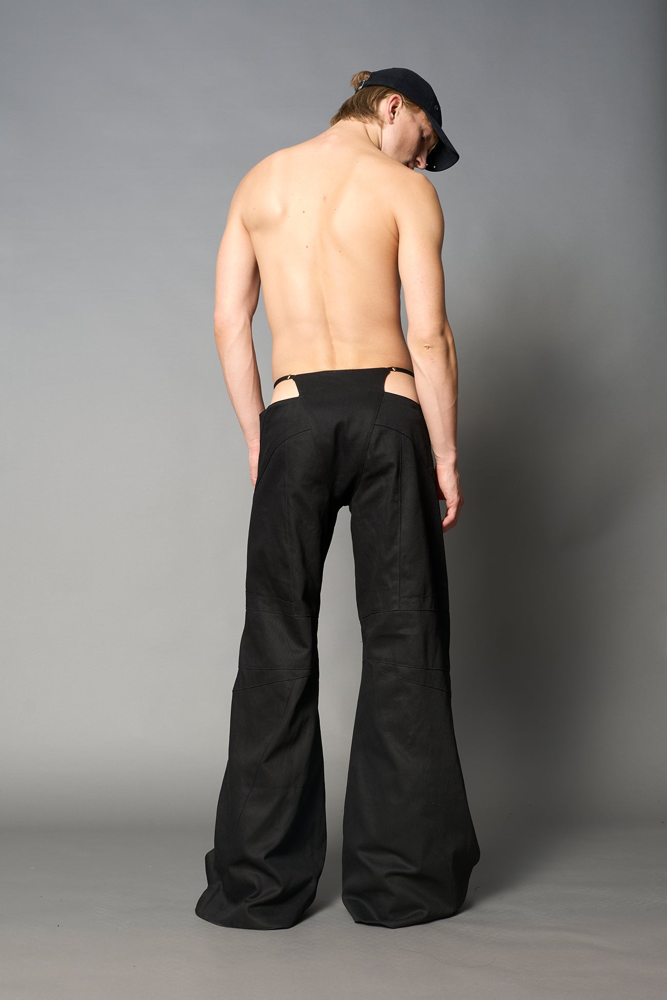 Low-rise flared pants in black cotton twill with bold side cut-outs and thong strap detailing. Featuring moto-style paneling, front zip closure, and a sculpted fit. A statement piece for rave, nightlife, and runway-inspired looks.