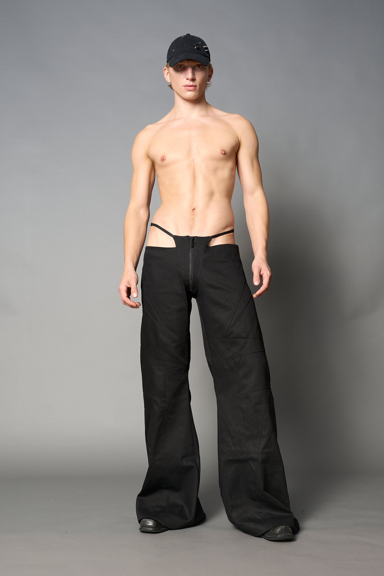 Low-rise flared pants in black cotton twill with bold side cut-outs and thong strap detailing. Featuring moto-style paneling, front zip closure, and a sculpted fit. A statement piece for rave, nightlife, and runway-inspired looks.