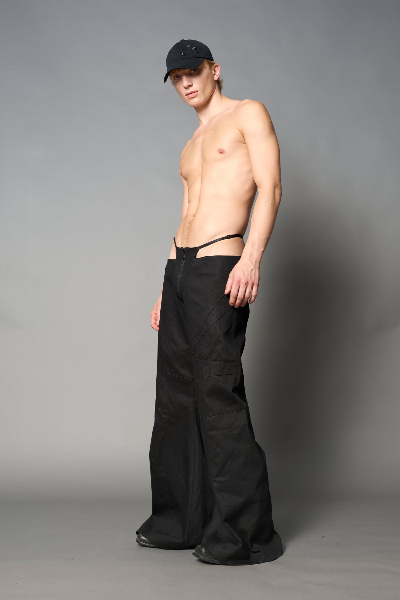 Low-rise flared pants in black cotton twill with bold side cut-outs and thong strap detailing. Featuring moto-style paneling, front zip closure, and a sculpted fit. A statement piece for rave, nightlife, and runway-inspired looks.