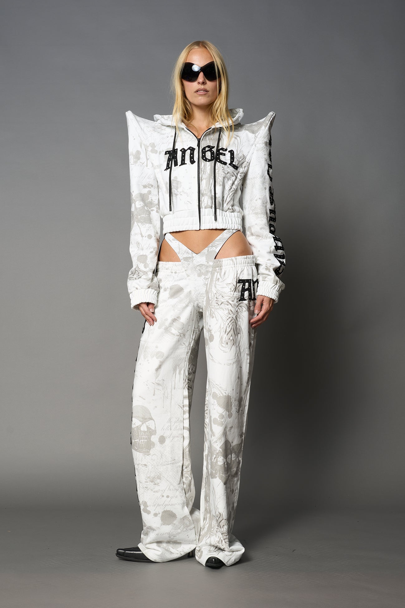 Model wearing low-rise white print wide-leg pants featuring a V-shaped waistband and detachable thong strap detail.