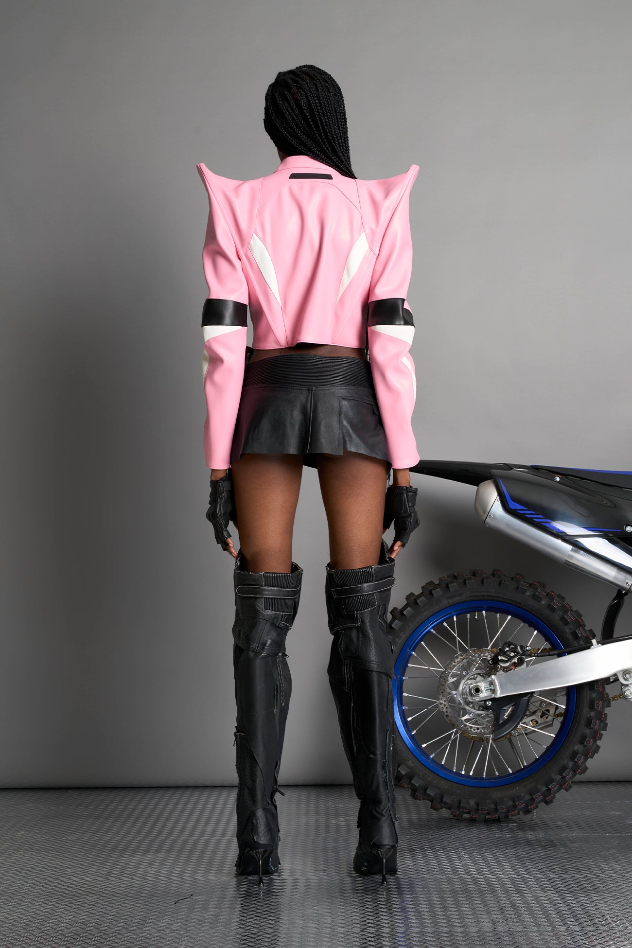 Model wears the pink Namilia Spike Moto Jacket with wire shoulder pads, branded zipper, band collar, zippered breast and waist pockets, motocross-style lines, and black logo rubber tag on the back.