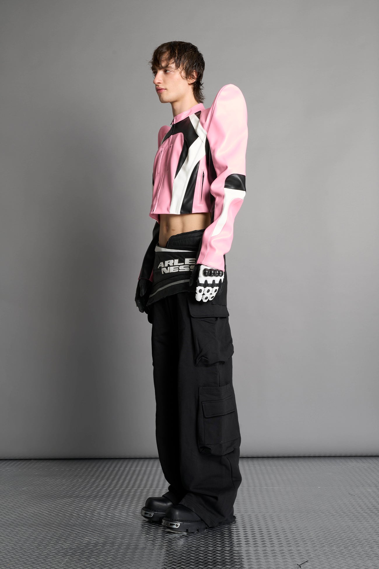 Model wears the pink Namilia Spike Moto Jacket with wire shoulder pads, branded zipper, band collar, zippered breast and waist pockets, motocross-style lines, and black logo rubber tag on the back.