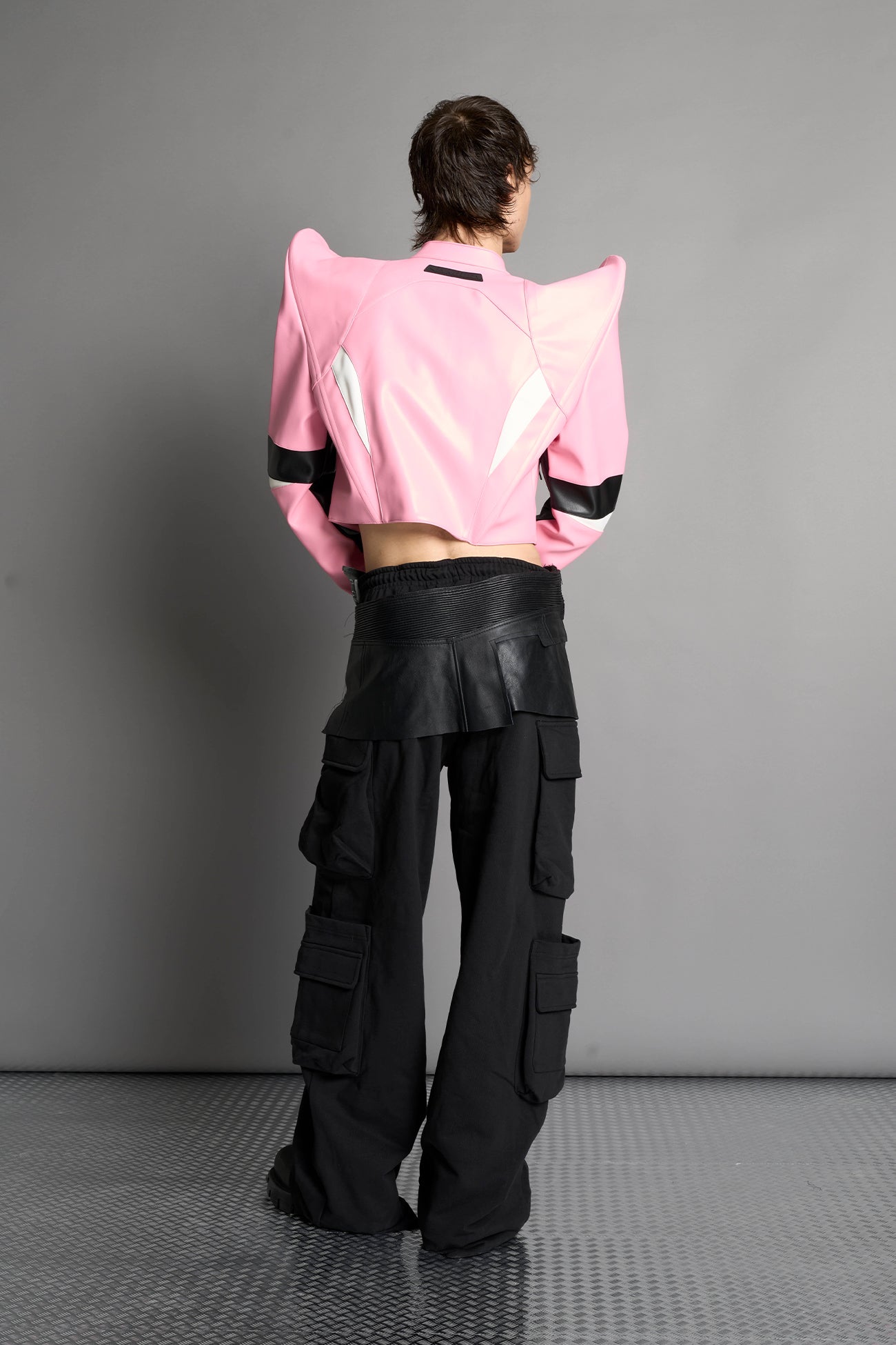 Model wears the pink Namilia Spike Moto Jacket with wire shoulder pads, branded zipper, band collar, zippered breast and waist pockets, motocross-style lines, and black logo rubber tag on the back.