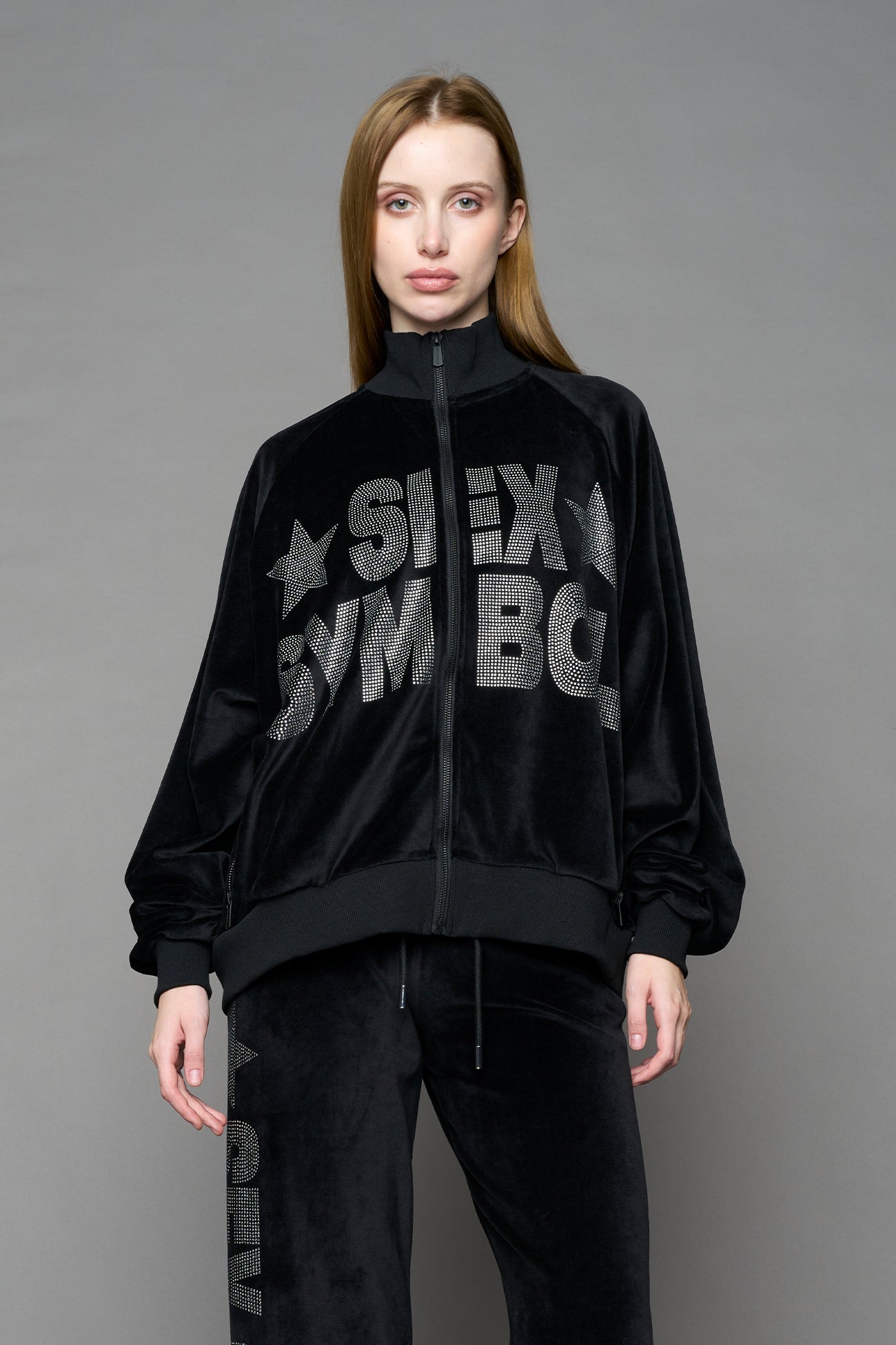 Person wearing a black velour jacket with 'S*X SYMBOL' crystal slogan on a gray background