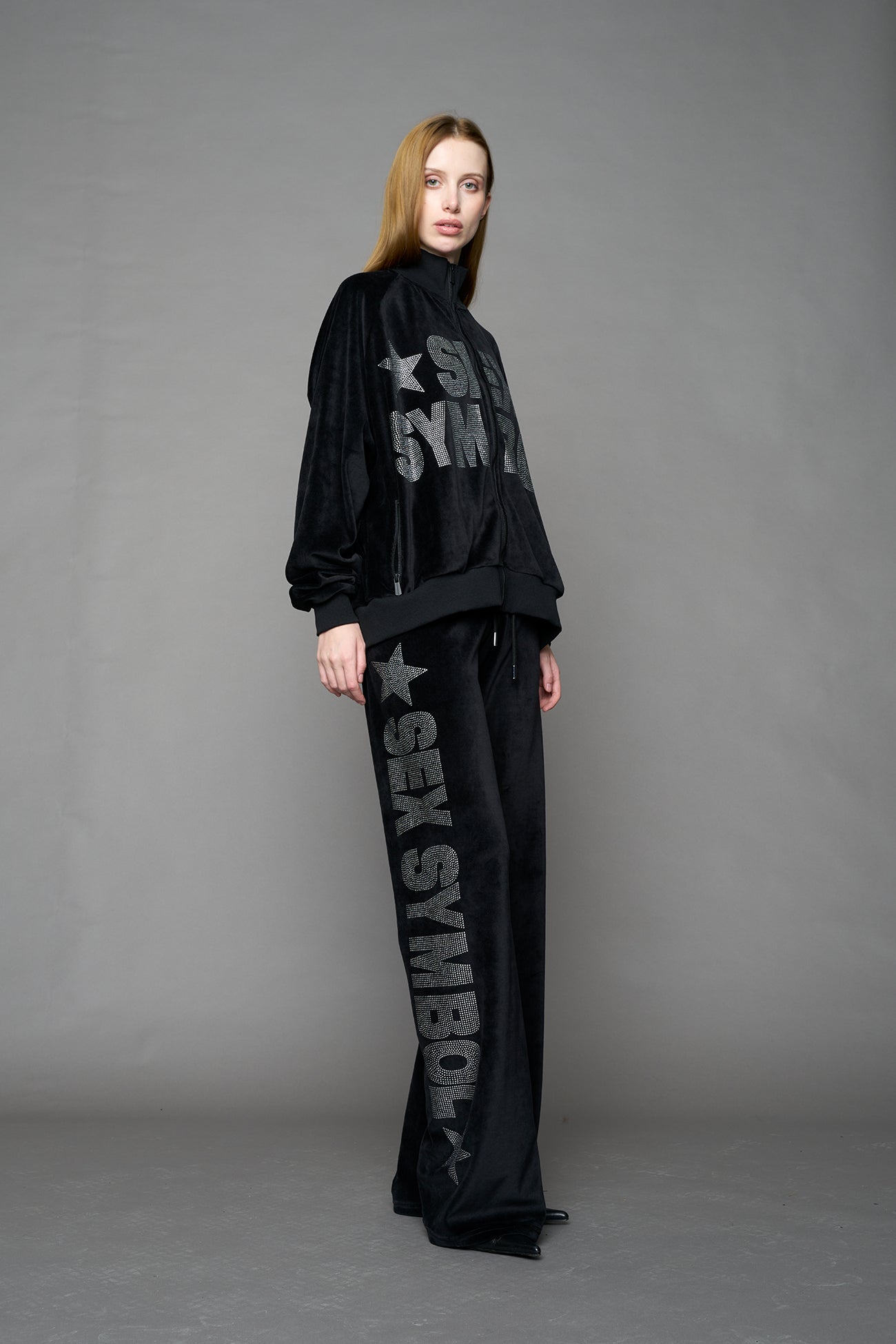 Person wearing a black velour jacket with 'S*X SYMBOL' crystal slogan on a gray background