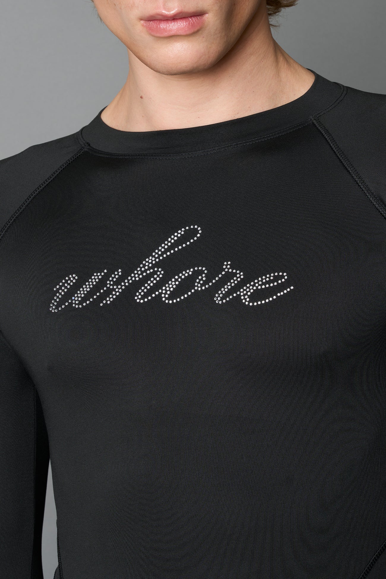 Male model wearing a black high-cut thong bodysuit with long sleeves and thumbhole details, edgy avant-garde fashion on a neutral grey studio background