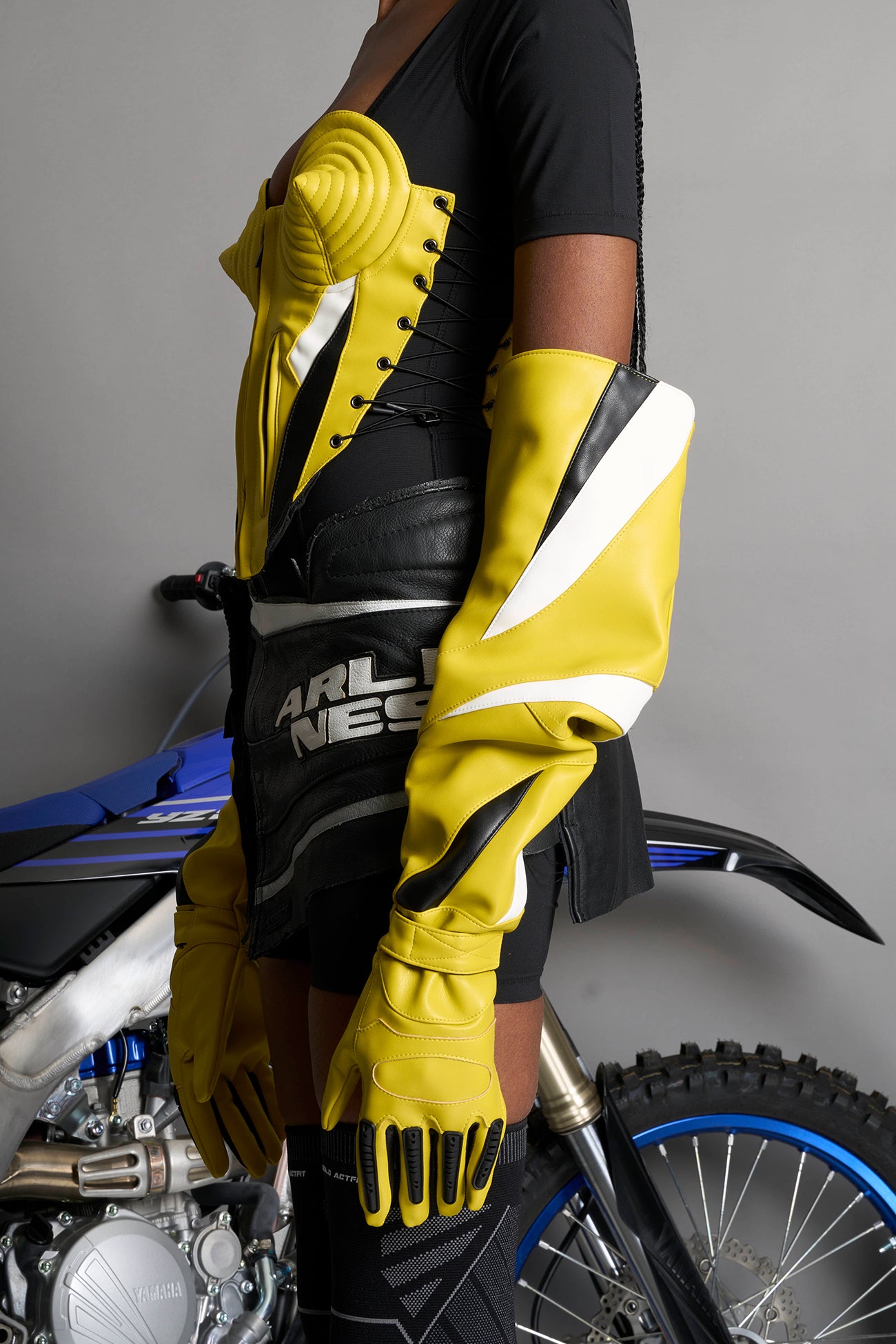 model wears yellow vegan leather elbow-length moto gloves with motocross-style lines, contrast panelling, rubber finger protection, lycra webbing, and velcro wrist fasteners.
