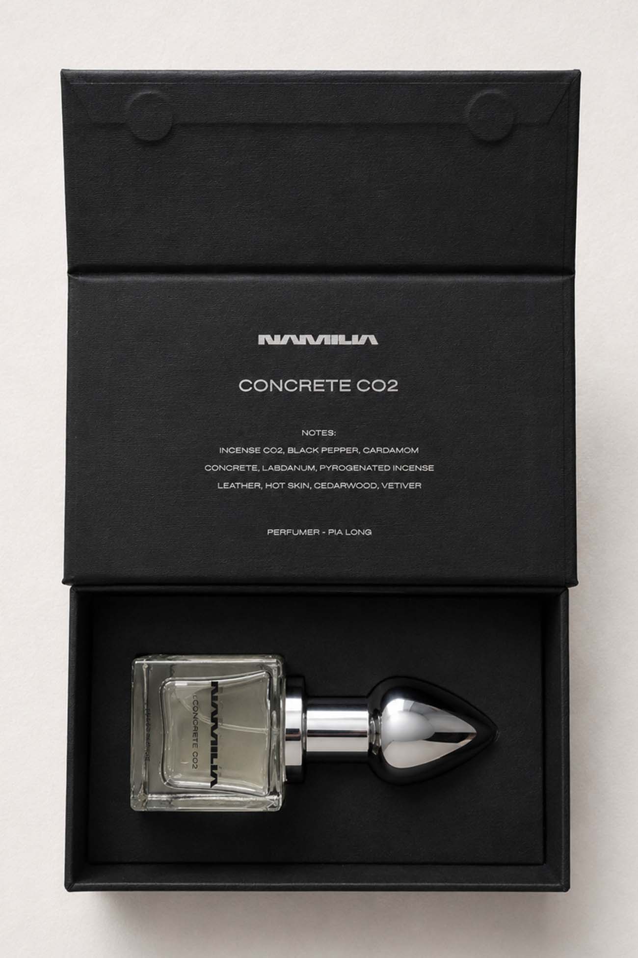 A bottle of Namila Concrete CO2 Eau de Parfum with a reflective silver plug-shaped cap and a clear glass base showing a pale yellow liquid, set against a solid black background.