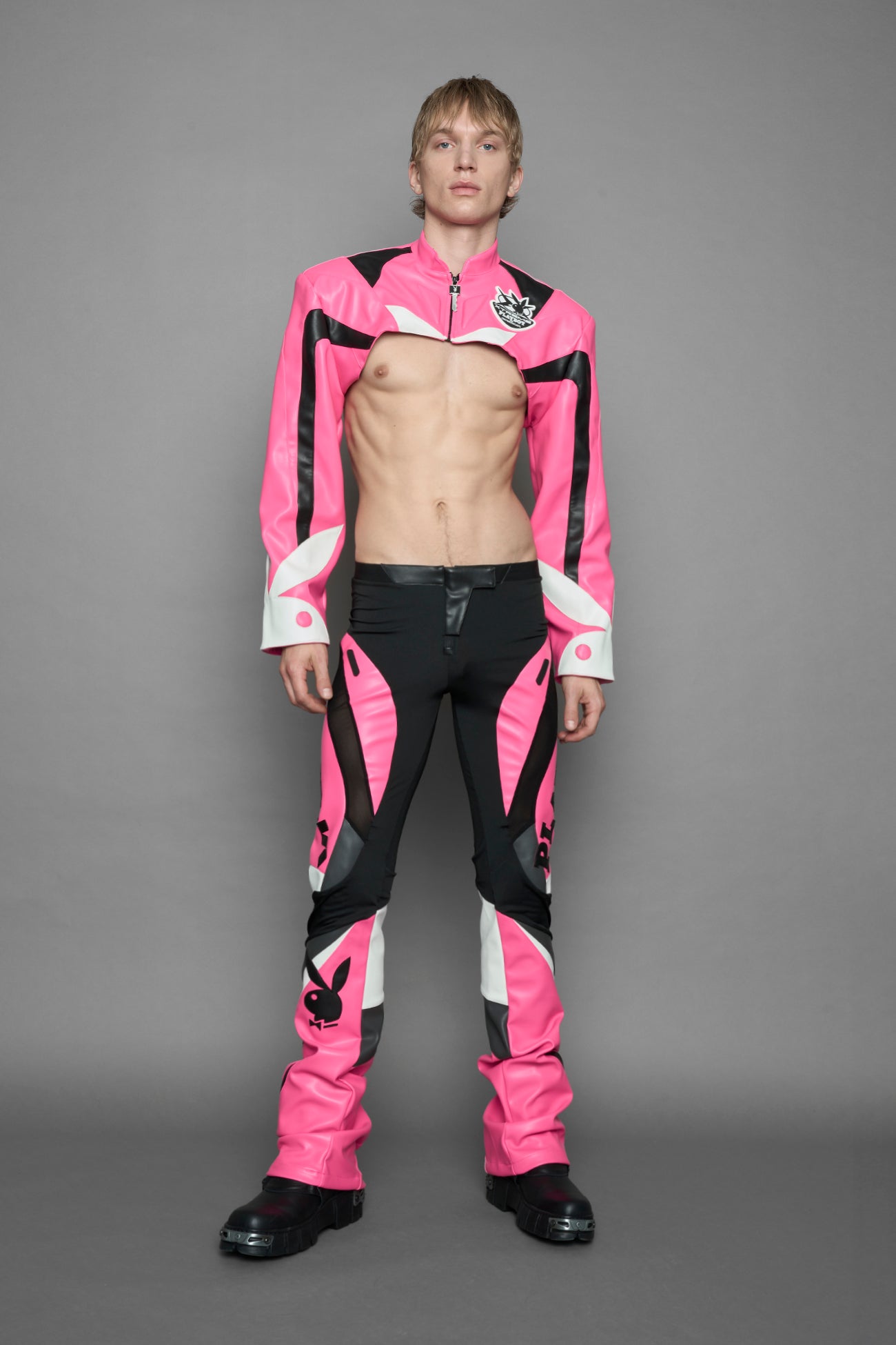 model wearing a vegan leather cropped jacket with broad shoulders in pink with black and white paneling. The jacket has PU leather embroidery of 'Namilia' and 'Playboy' with a key formed zipper pull and a rubbertag on the left breast.