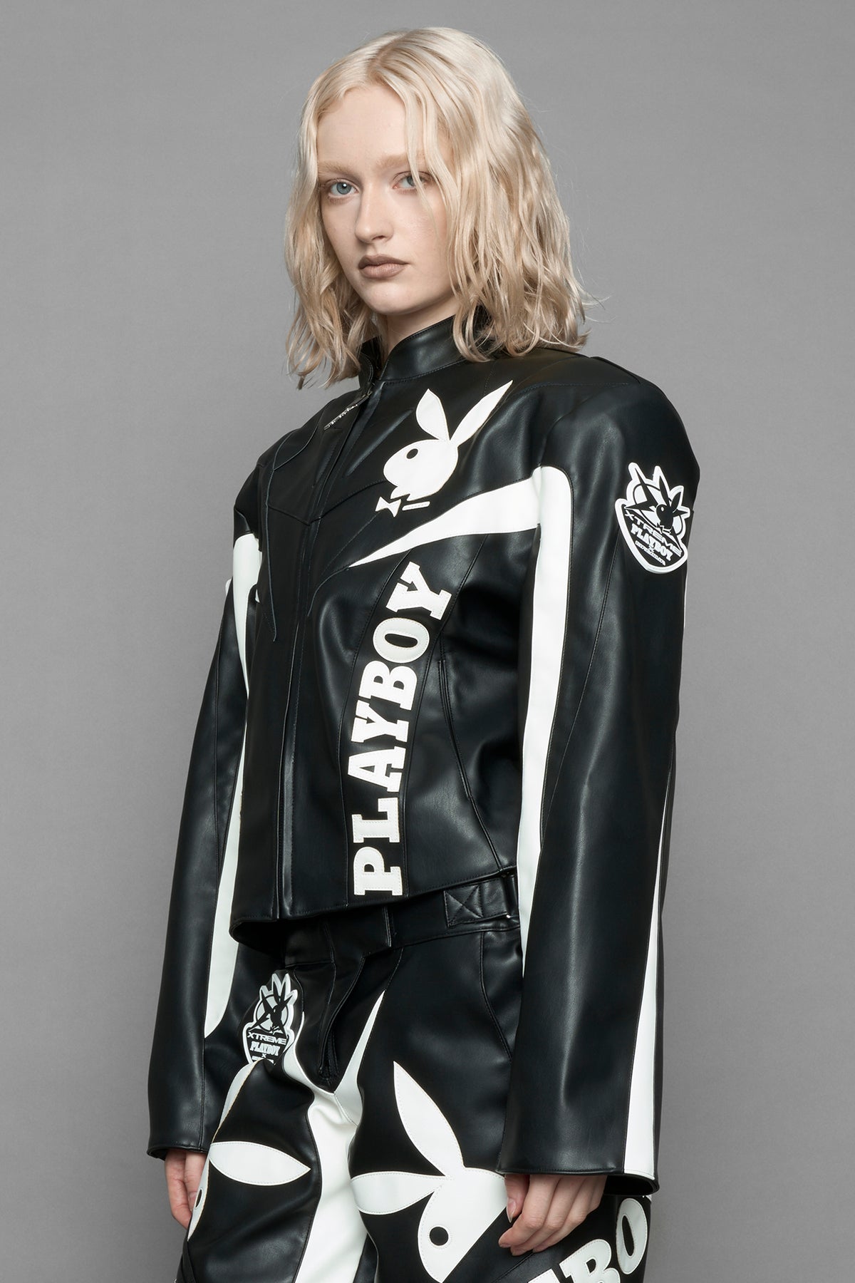 model wearing black vegan leather zip-up jacket with white PU leather 'Namilia' and 'Playboy' embroidery and black and white paneling. On the left chest there is a white Playboy Bunny Embroidery.