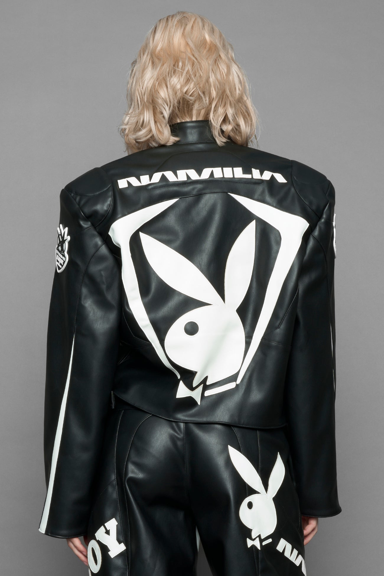 model wearing black vegan leather zip-up jacket with white PU leather 'Namilia' and 'Playboy' embroidery and black and white paneling. On the left chest there is a white Playboy Bunny Embroidery.