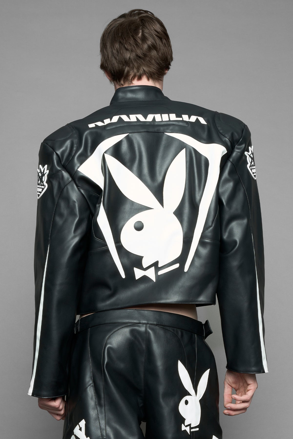 model wearing black vegan leather zip-up jacket with white PU leather 'Namilia' and 'Playboy' embroidery and black and white paneling. On the left chest there is a white Playboy Bunny Embroidery.