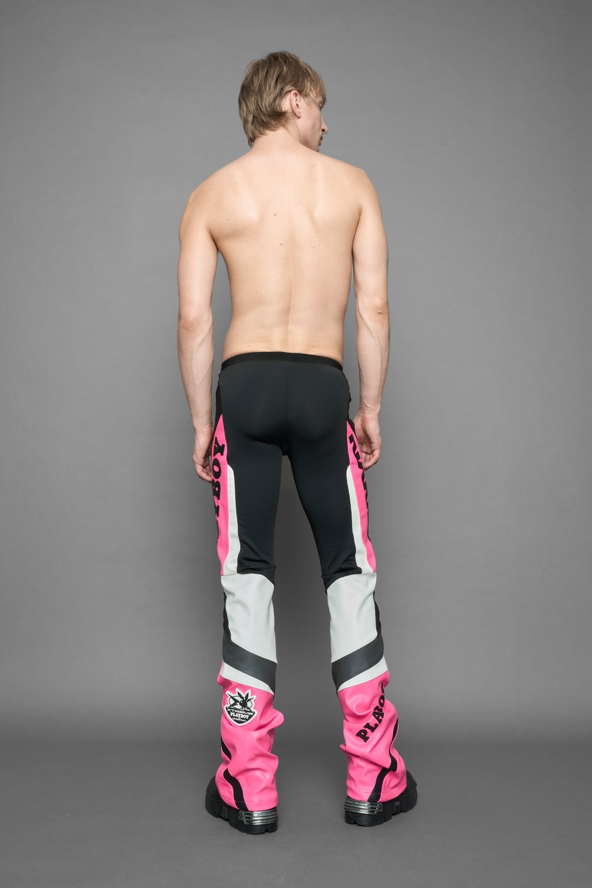 A female model wearing a pink and black form fitted Pants with PU Leather Embroidery of 'Namilia', 'Playboy'. The pants have black Mesh fabric and dark grey PU leather panels and are slightly flared from knee to hem.