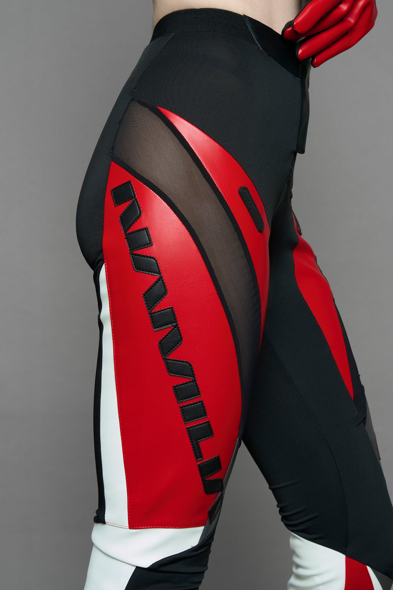model wearing a red and black form fitted Pants with PU Leather Embroidery of 'Namilia', 'Playboy'. The pants have black Mesh fabric and dark grey PU leather panels and are slightly flared from knee to hem