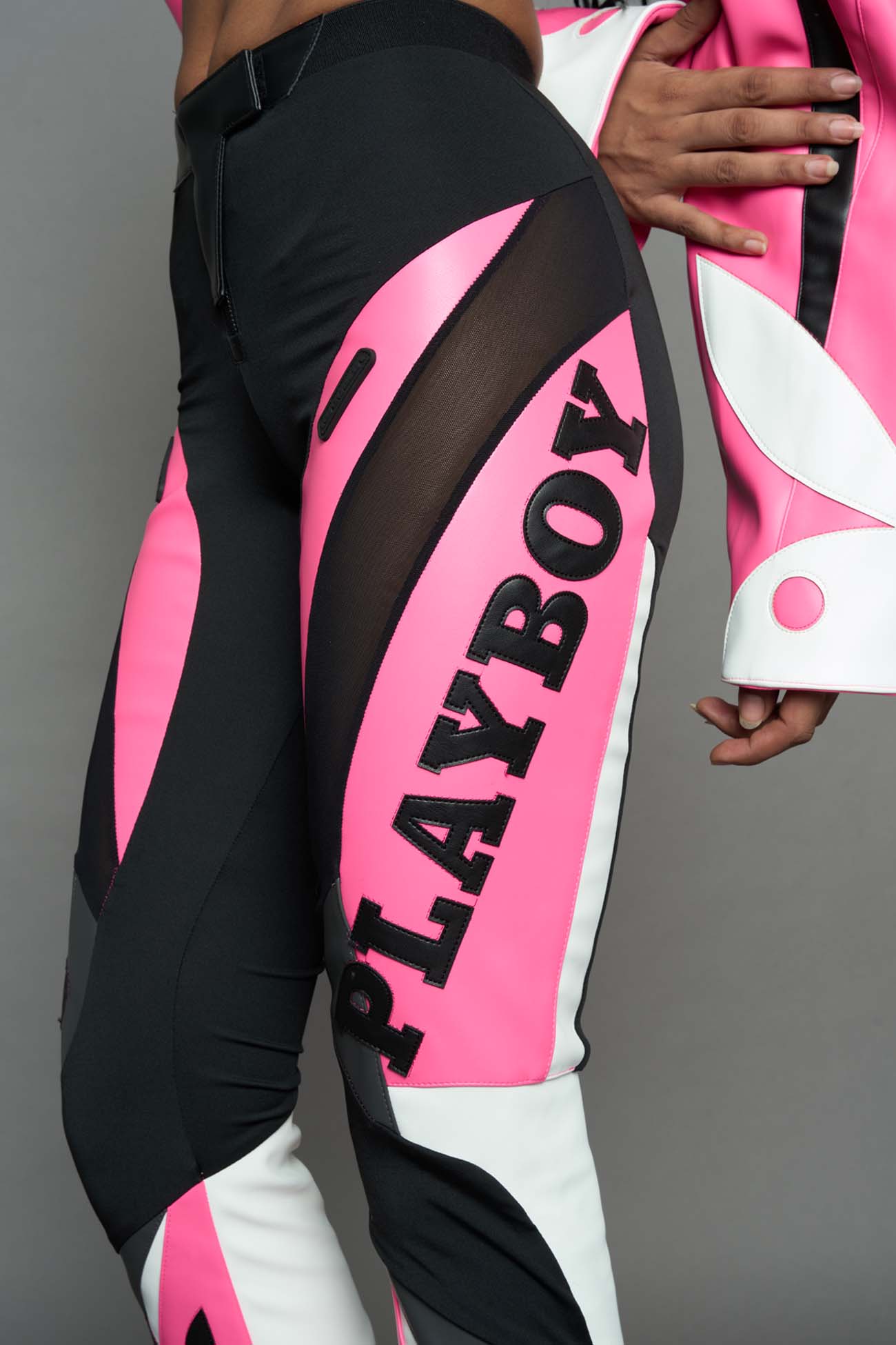 A female model wearing a pink and black form fitted Pants with PU Leather Embroidery of 'Namilia', 'Playboy'. The pants have black Mesh fabric and dark grey PU leather panels and are slightly flared from knee to hem.