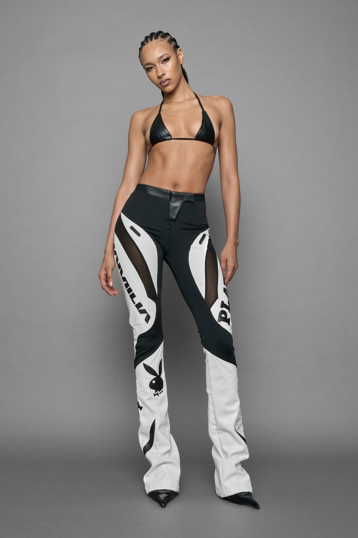 A female model wearing a white and black form fitted Pants with PU Leather Embroidery of 'Namilia', 'Playboy'. The pants have black Mesh fabric panels and are slightly flared from knee to hem