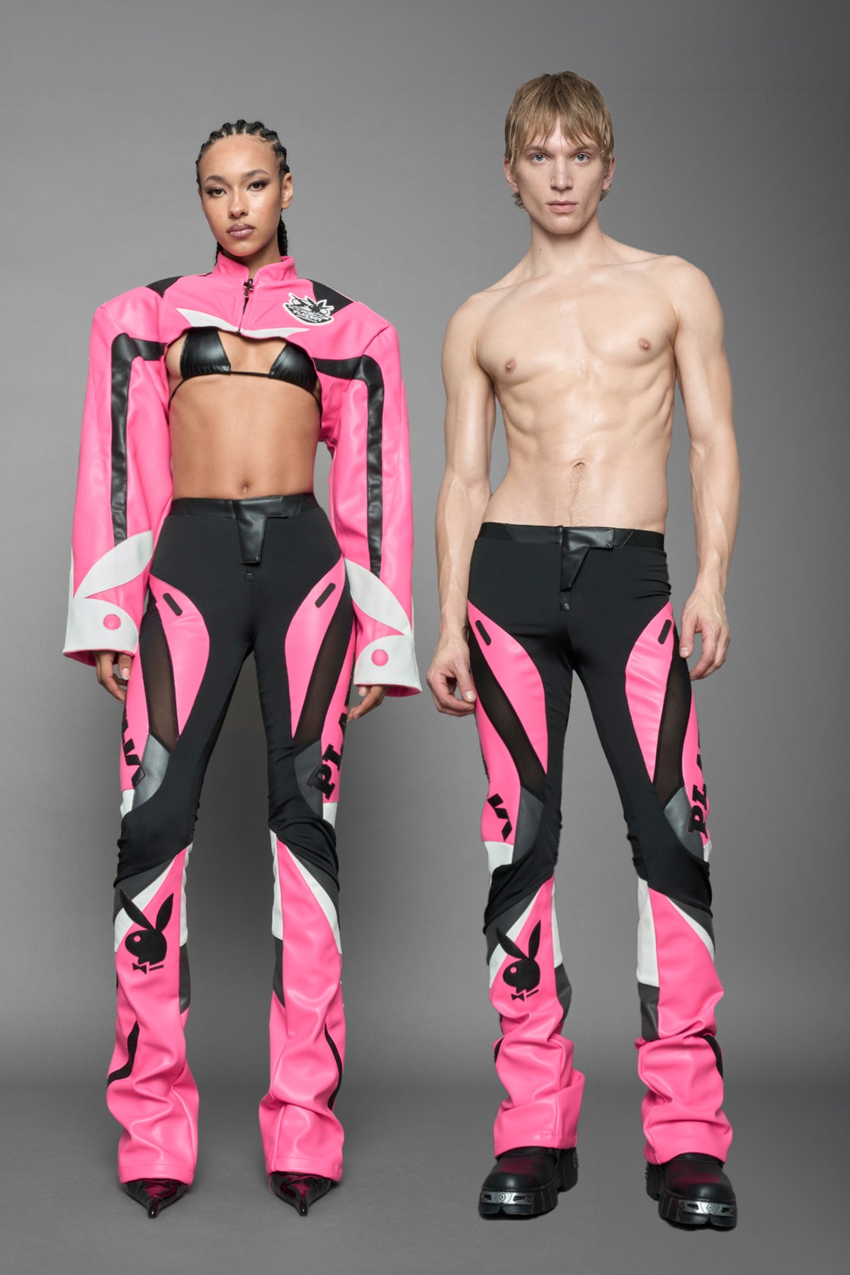 model wearing a pink and black form fitted Pants with PU Leather Embroidery of 'Namilia', 'Playboy'. The pants have black Mesh fabric and dark grey PU leather panels and are slightly flared from knee to hem.
