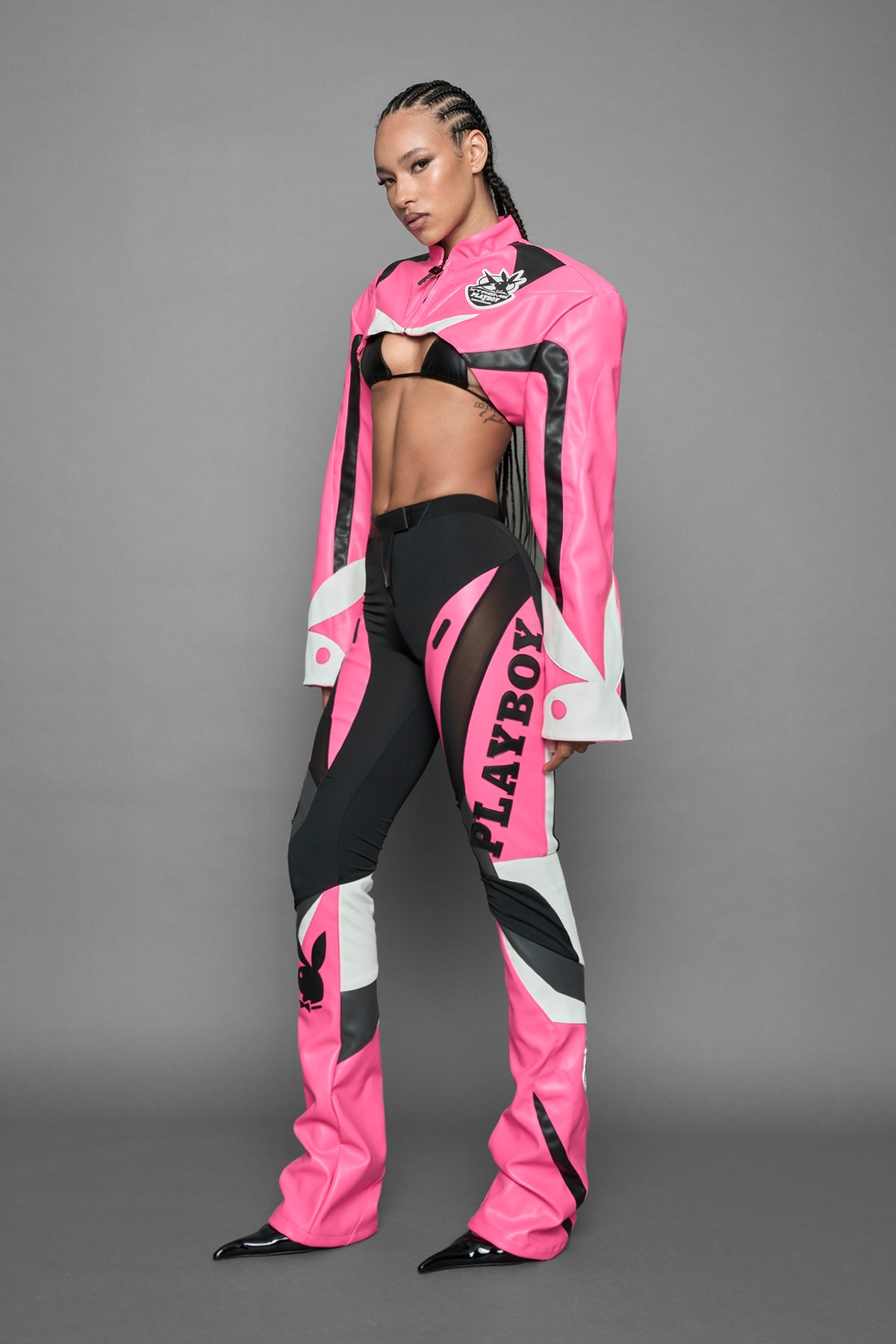 A female model wearing a pink and black form fitted Pants with PU Leather Embroidery of 'Namilia', 'Playboy'. The pants have black Mesh fabric and dark grey PU leather panels and are slightly flared from knee to hem.