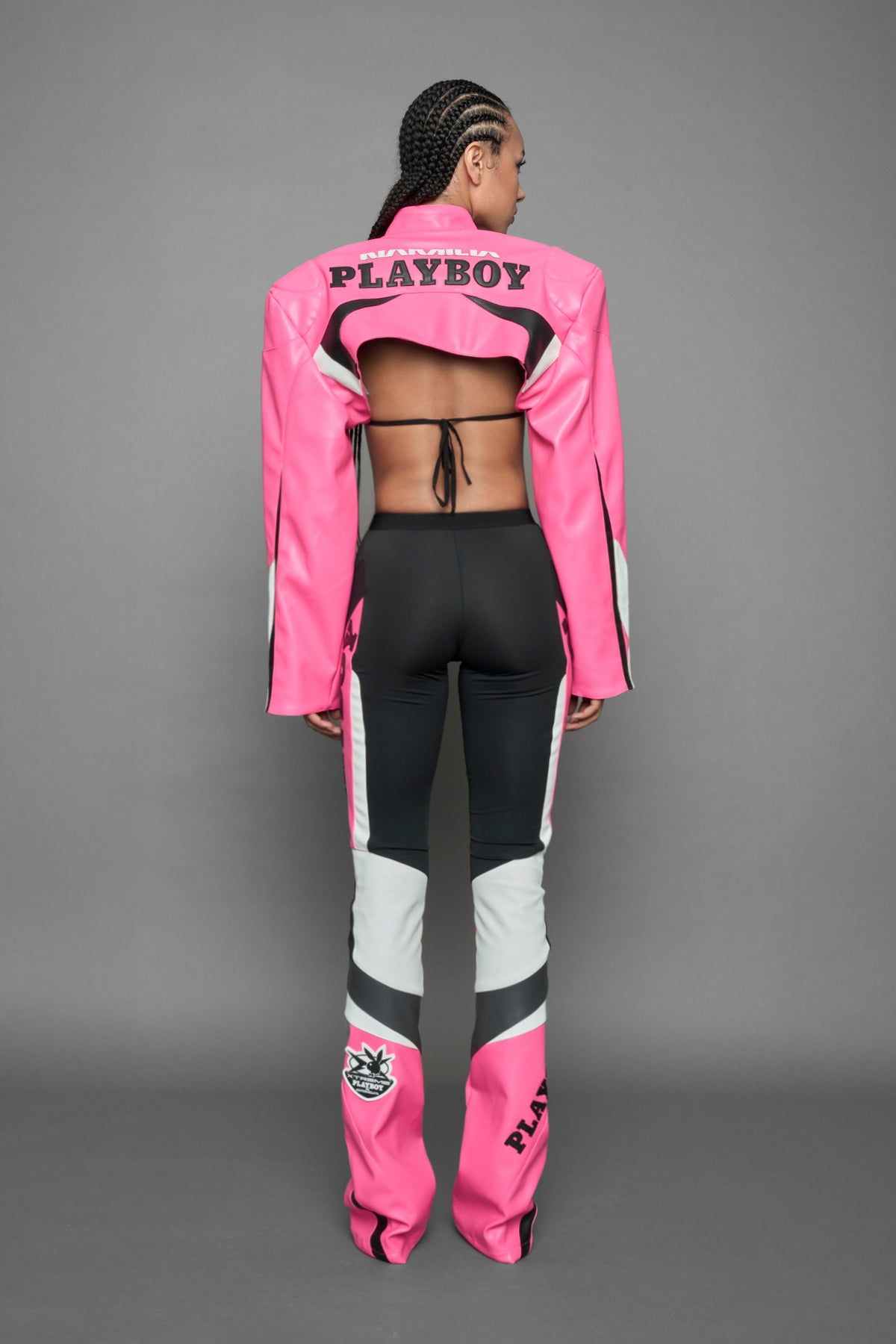 A female model wearing a pink and black form fitted Pants with PU Leather Embroidery of 'Namilia', 'Playboy'. The pants have black Mesh fabric and dark grey PU leather panels and are slightly flared from knee to hem.