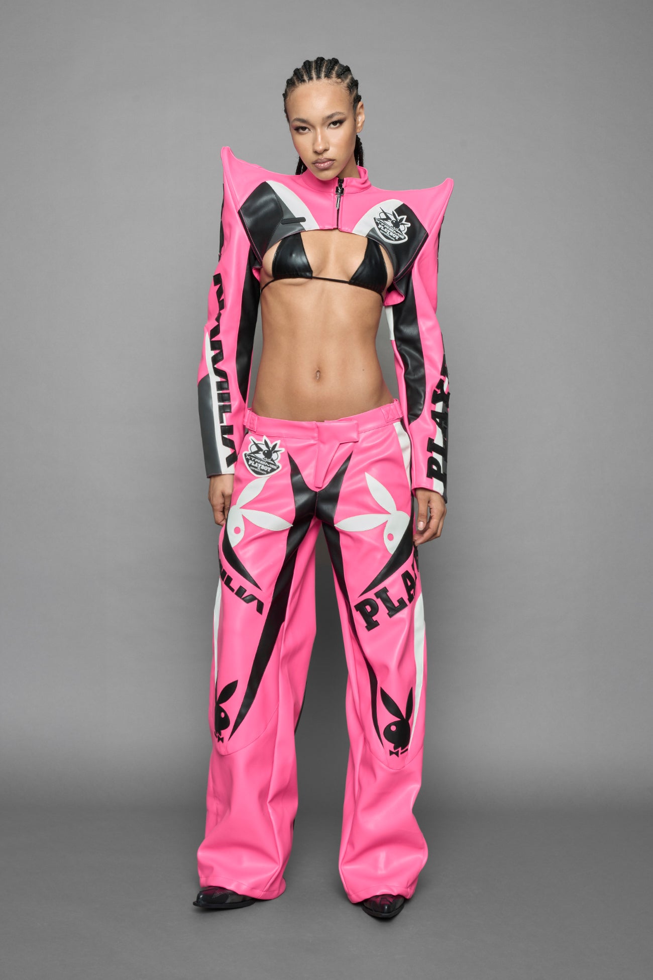 model wearing a vegan leather cropped jacket with spike silhouette shoulders in pink with black and white paneling. The jacket has PU leather embroidery of 'Namilia' and 'Playboy' with a key formed zipper pull and a rubbertag on the left breast.
