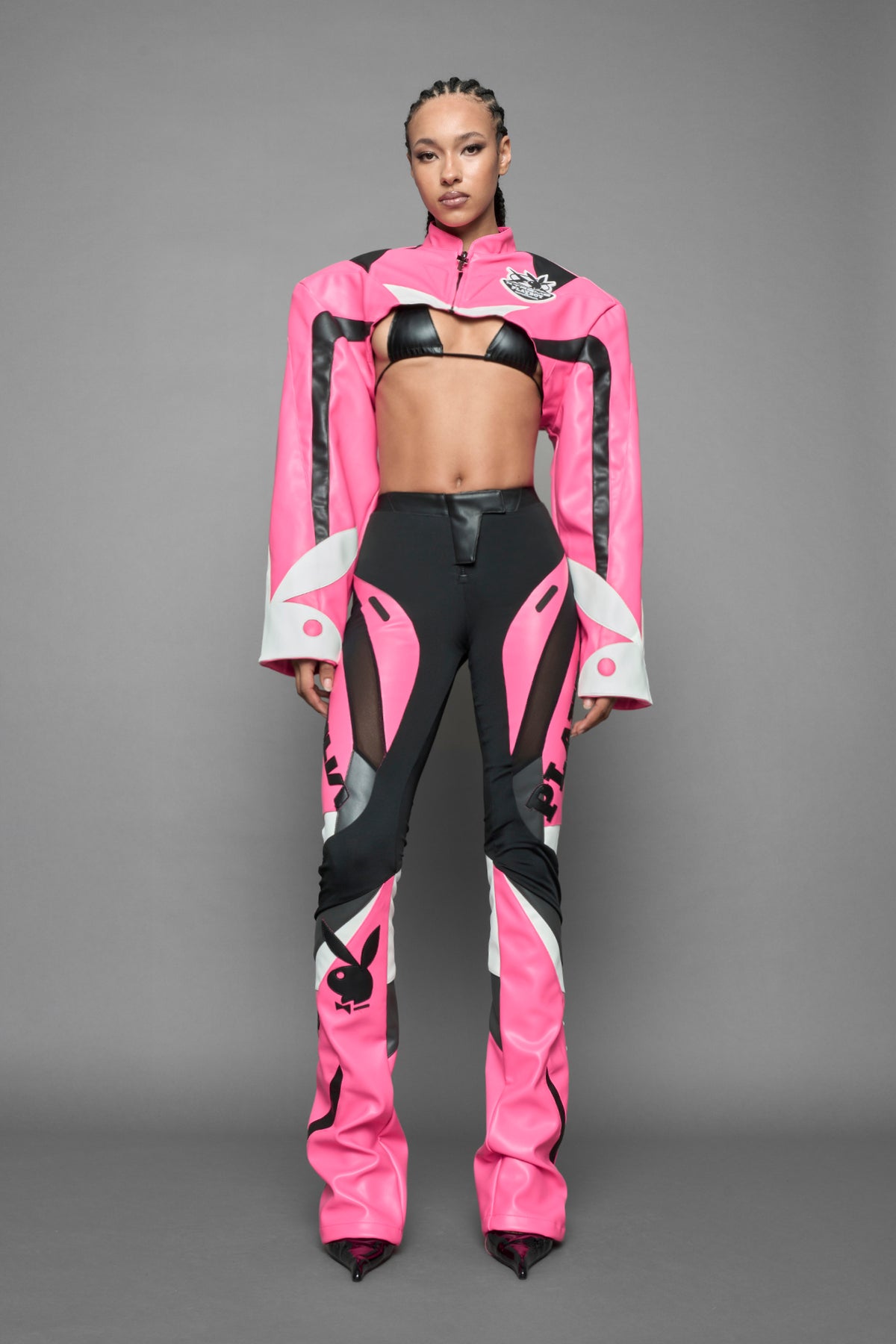 model wearing a vegan leather cropped jacket with broad shoulders in pink with black and white paneling. The jacket has PU leather embroidery of 'Namilia' and 'Playboy' with a key formed zipper pull and a rubbertag on the left breast.