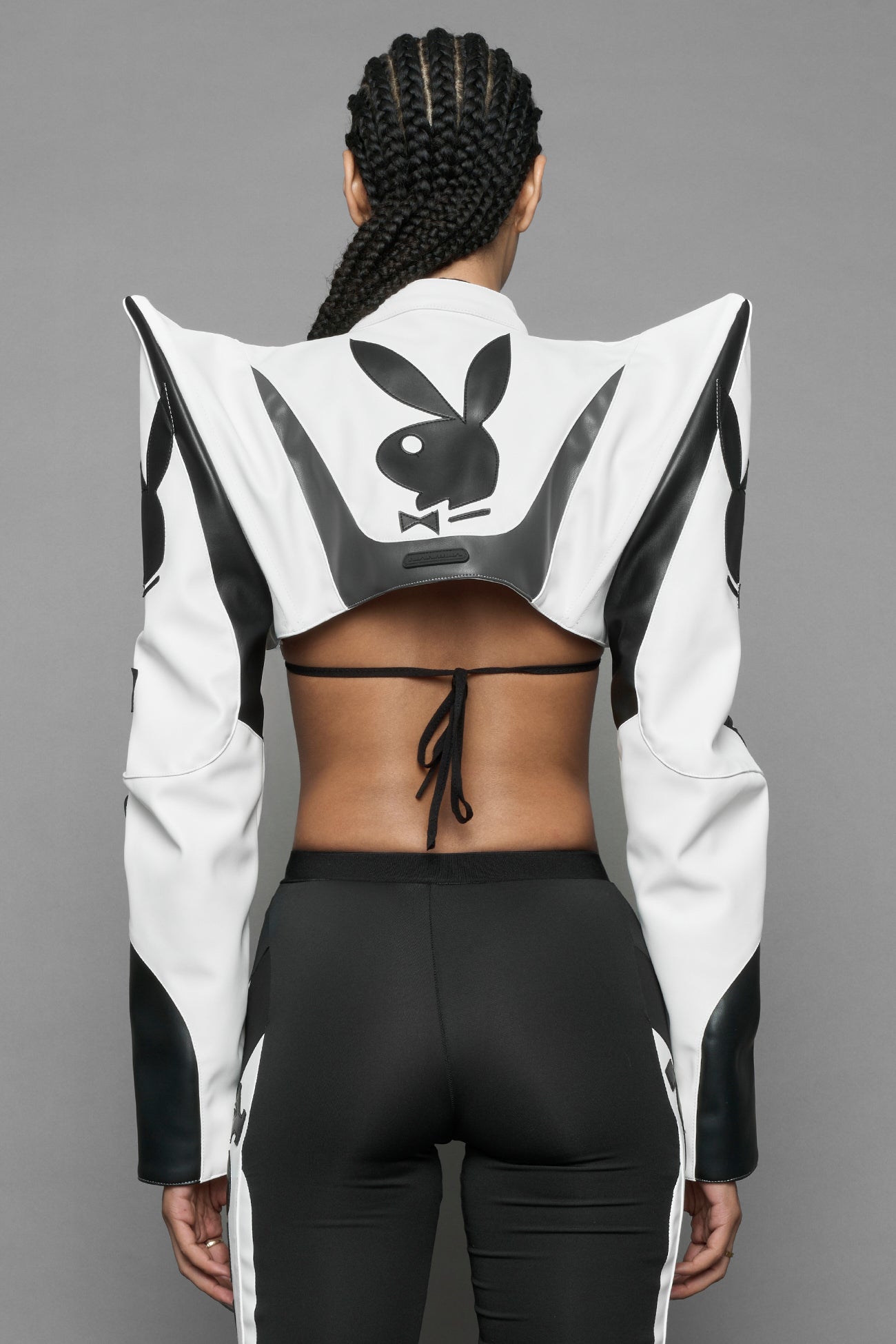 A female model wearing a vegan leather cropped jacket with spike silhouette shoulders in white with black paneling. The jacket has PU leather embroidery of 'Namilia' and 'Playboy' with a key formed zipper pull and a rubbertag on the left breast.