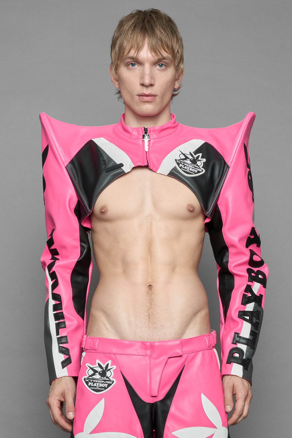 A male model wearing a vegan leather cropped jacket with spike silhouette shoulders in pink with black and white paneling. The jacket has PU leather embroidery of 'Namilia' and 'Playboy' with a key formed zipper pull and a rubbertag on the left breast.