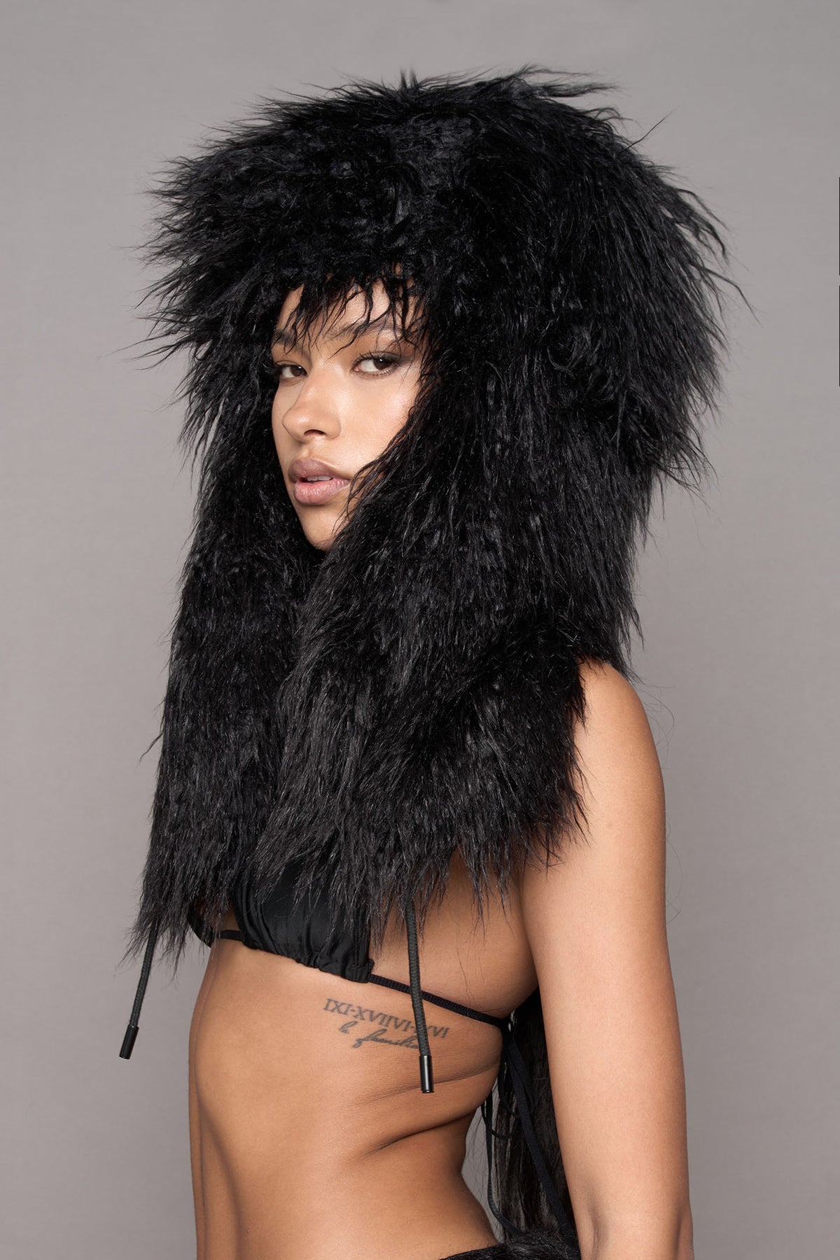A close up of amodel wearing a voluminous black faux fur hat with earflaps and cords, styled over a black bikini top against a solid gray studio background.