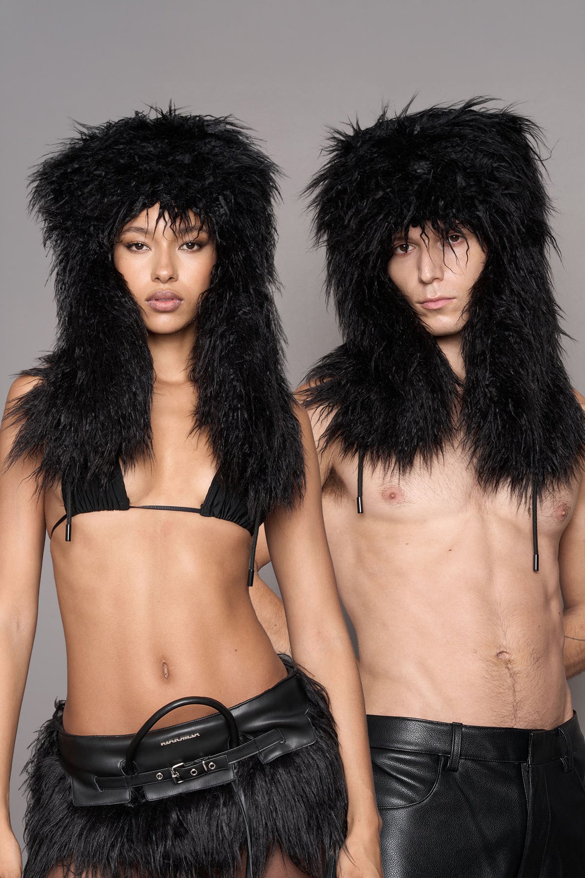 Two models wearing matching black faux fur oversized hat with earflaps and cords, styled with a matching black faux-leather mini skirt a black bikini top, posed against a solid gray studio background.