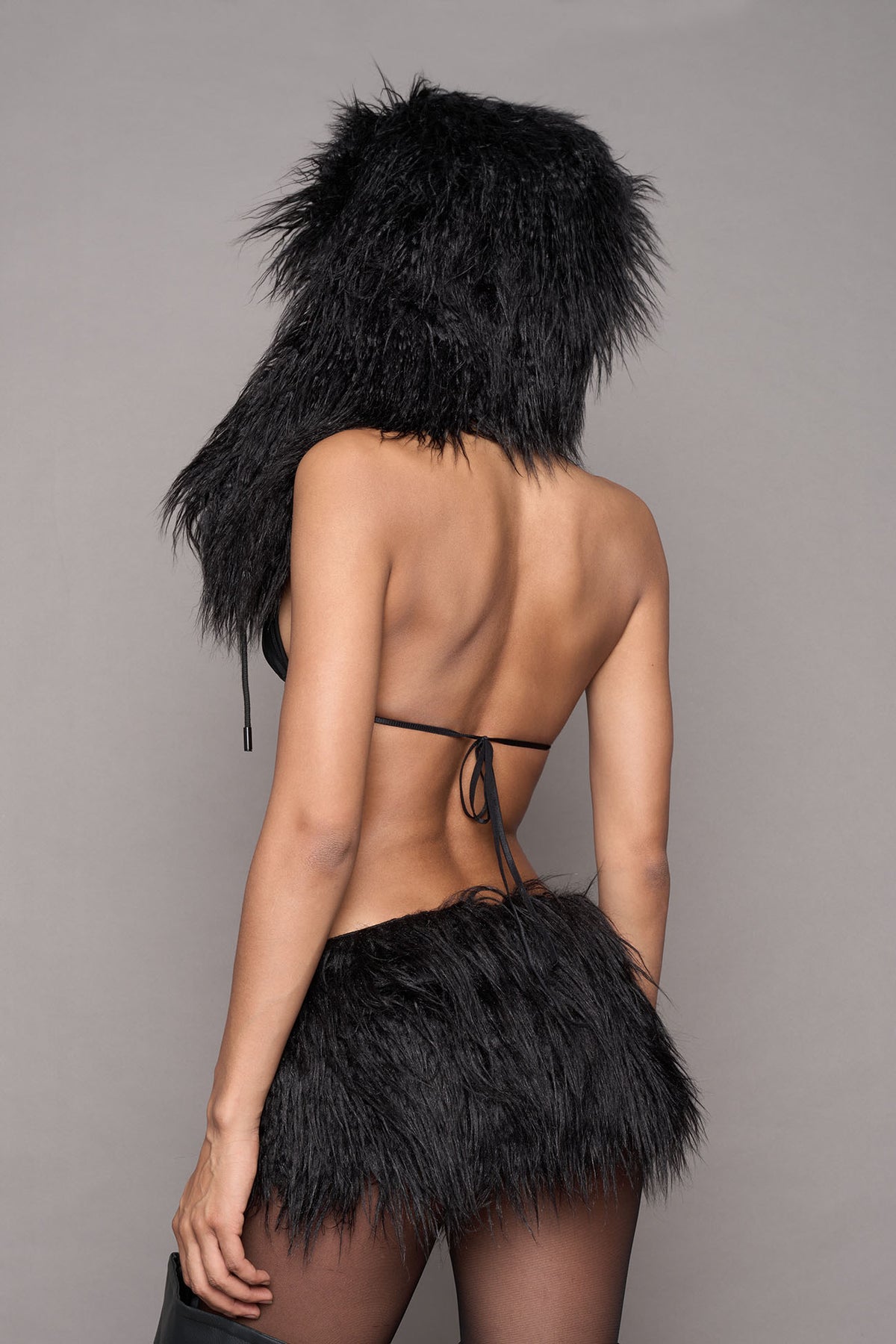 A model shown from the back wearing a black faux fur hat with earflaps and cords styled with a black string bikini top, and a black matching skirt photographed against a solid gray studio background.
