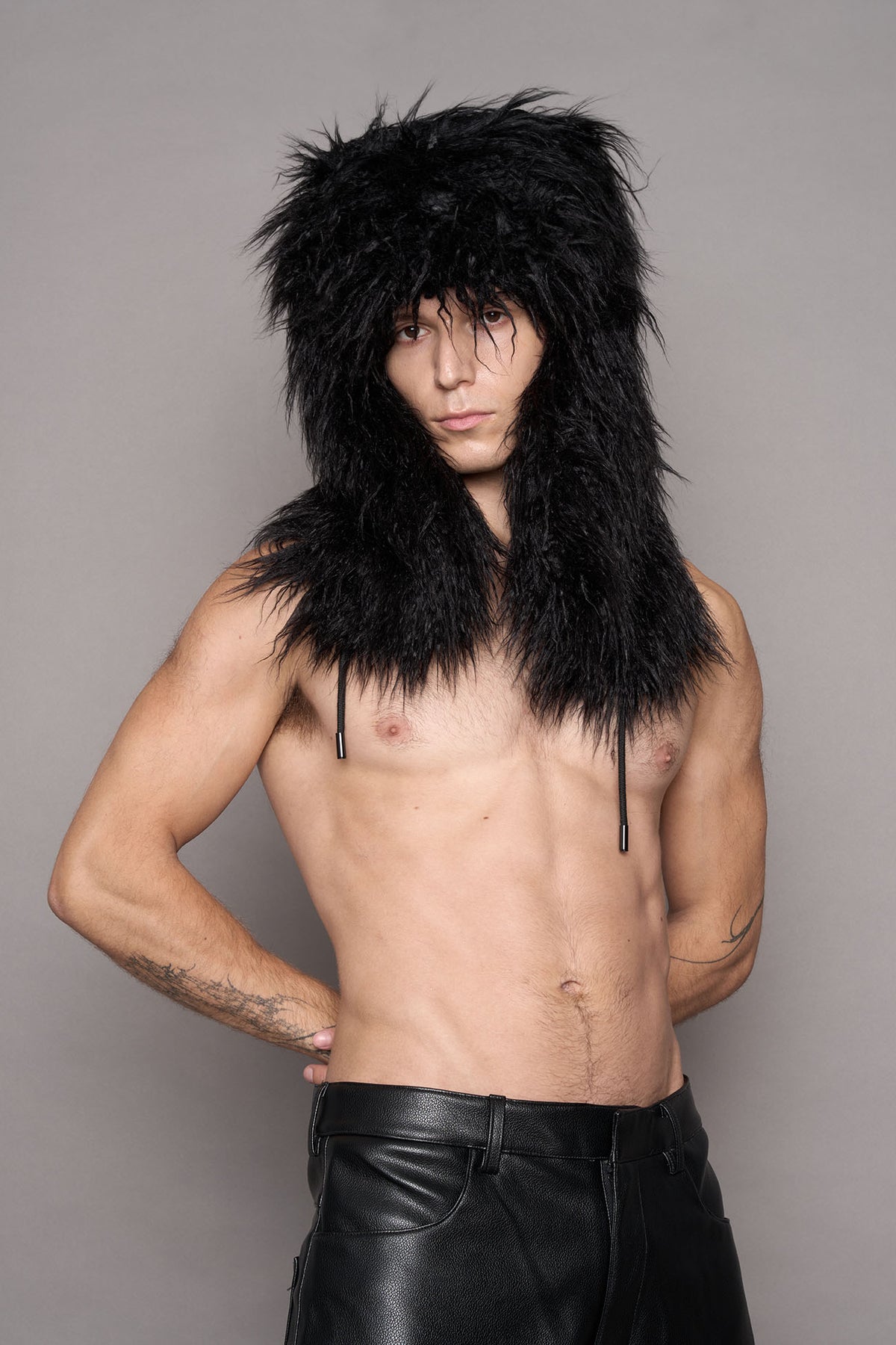 A shirtless man wearing a voluminous black faux fur oversized hat posed against a plain gray studio background.