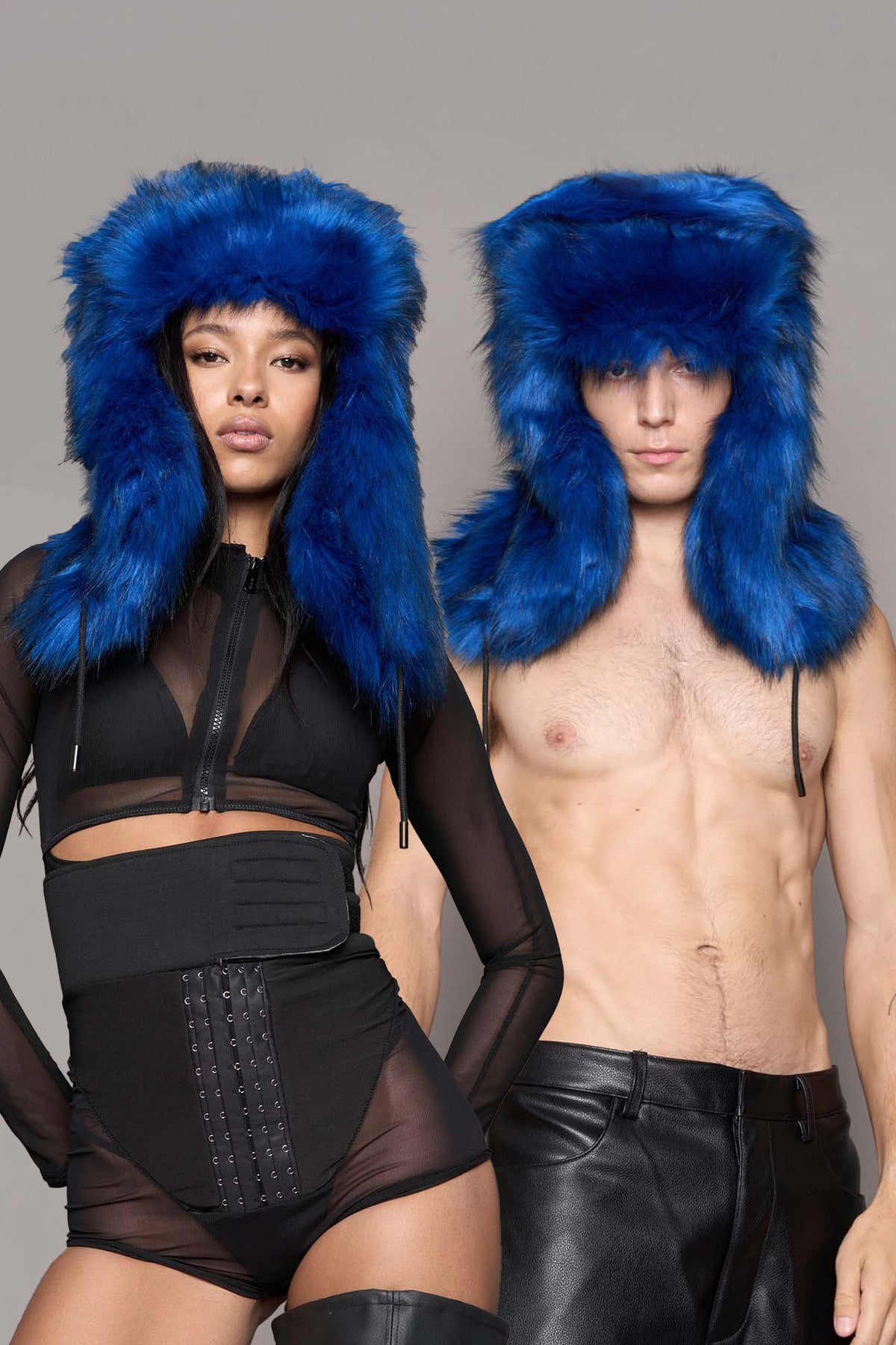 A man and a woman modeling matching bright blue faux-fur ushanka-style hats against a plain light gray studio background. The woman wears a black sheer outfit, and the man wears black pants.