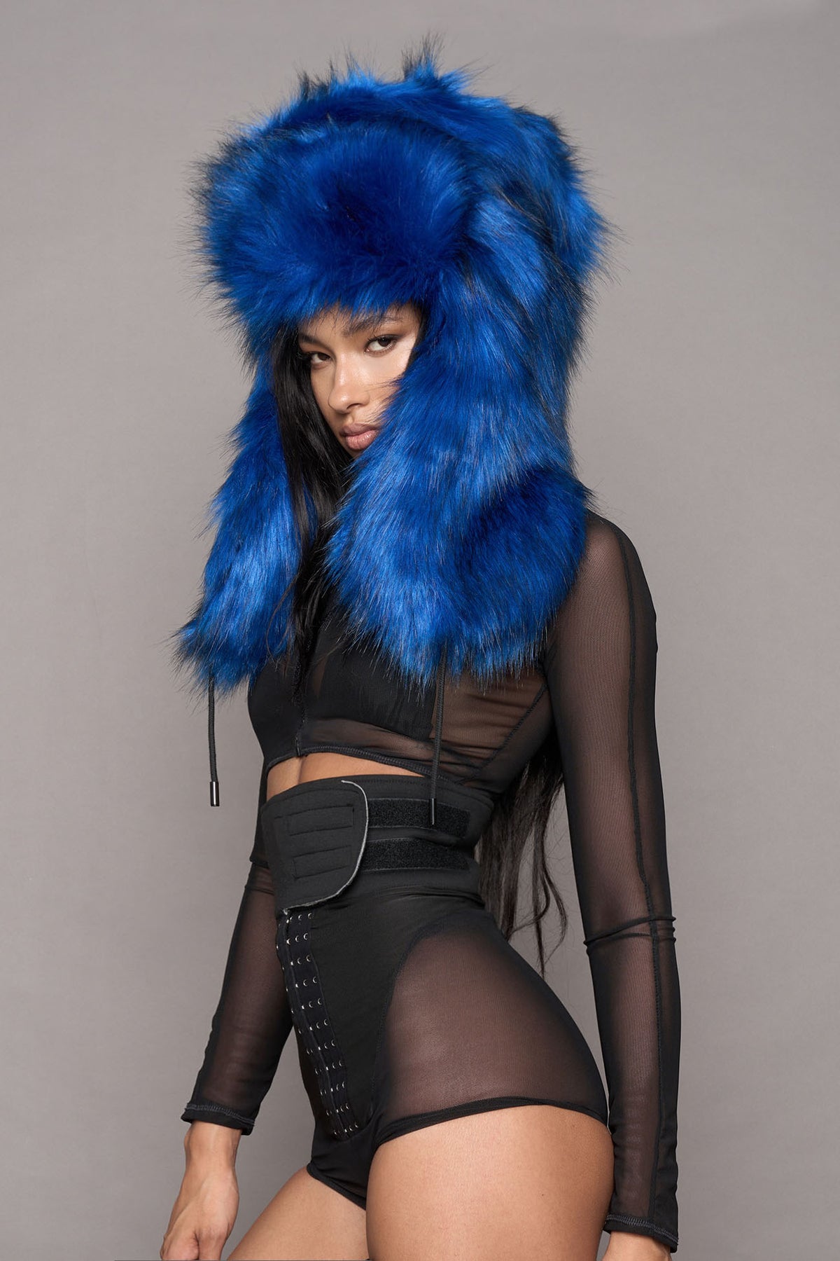 A model wearing a vibrant blue faux-fur hood with black trim, paired with a black sheer long-sleeve crop top, matching black corset-style shorts, and black thigh-high boots, posed against a solid gray studio background.