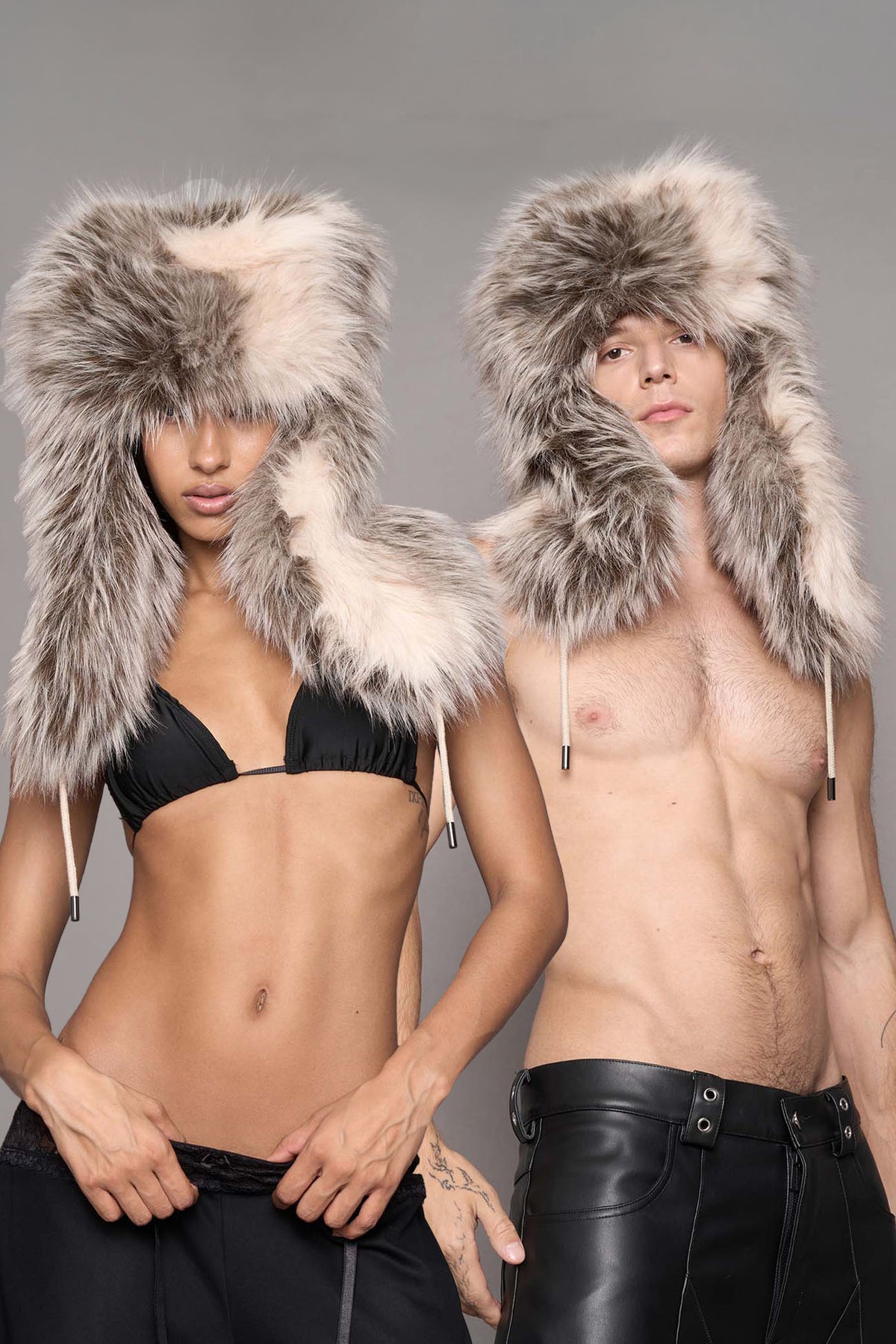 Two models wearing oversized faux-fur ushanka-style hooded hats with drawstrings, shown against a plain gray studio background.