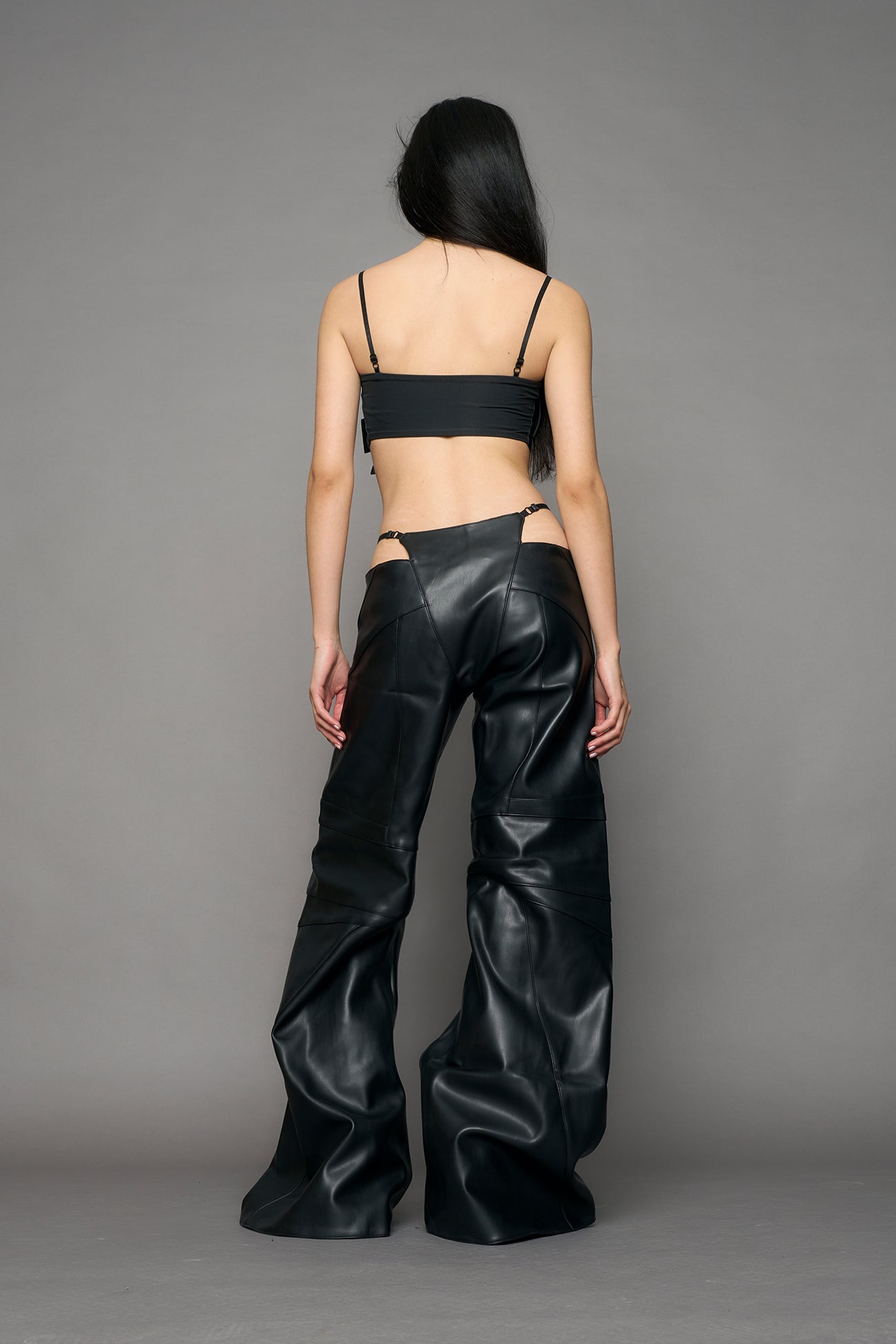 Low-rise flared pants in black faux leather with bold side cut-outs and thong strap detailing. Featuring moto-style paneling, front zip closure, and a sculpted fit. A statement piece for rave, nightlife, and runway-inspired looks.