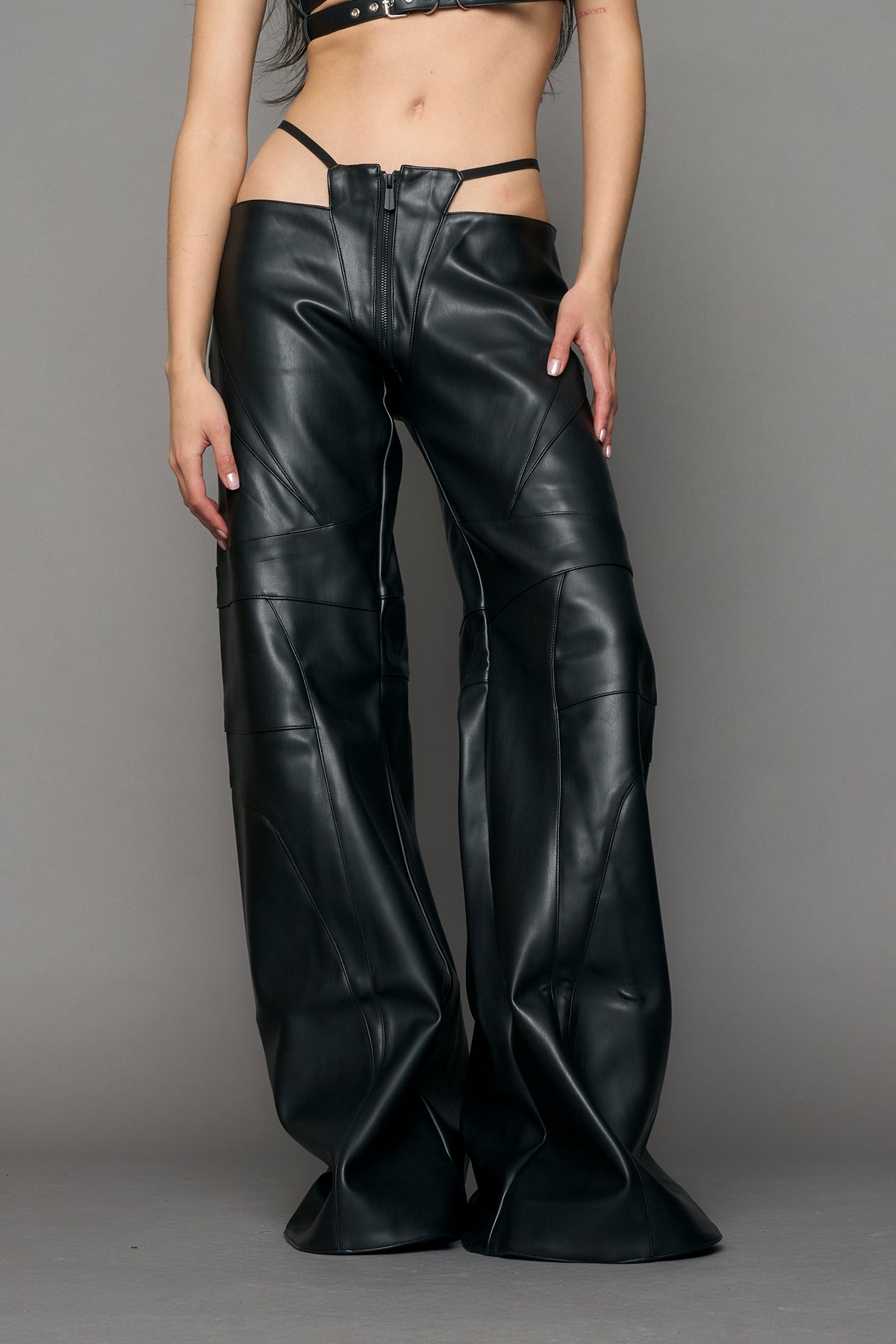 Low-rise flared pants in black faux leather with bold side cut-outs and thong strap detailing. Featuring moto-style paneling, front zip closure, and a sculpted fit. A statement piece for rave, nightlife, and runway-inspired looks.