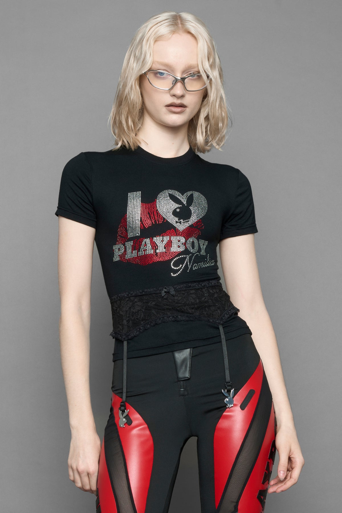 A female model wearing a black short-sleeve graphic t-shirt with rhinestone embroidery of red lip print and silver 'I <heart> PLAYBOY' design. T-Shirt has a lingerie black lace garter with suspender with metal silver Bunny Heads attached.